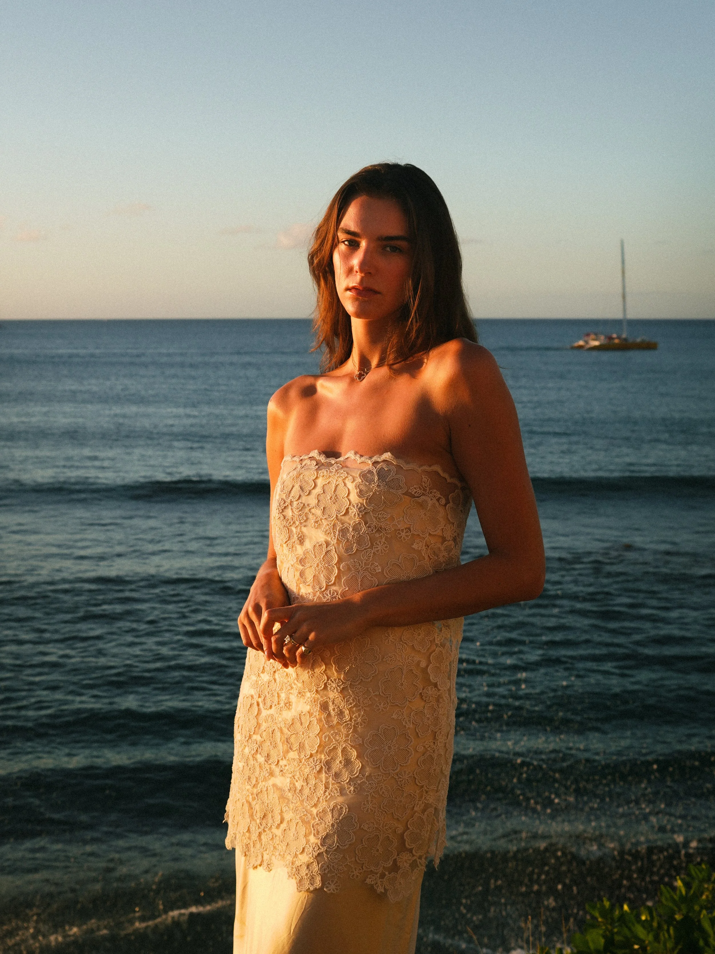 A woman in a strapless lace dress standing on a beach during sunset with the ocean and a sailboat in the background.