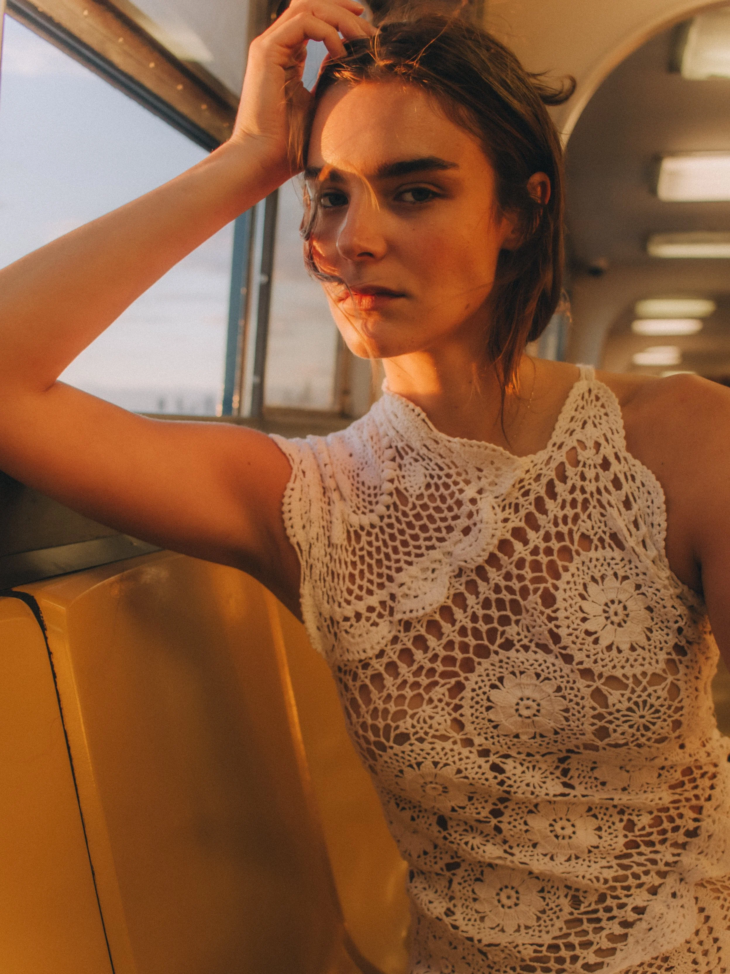 A woman with shoulder-length brown hair and light skin wearing a white crochet dress, sitting inside a yellow and black vehicle, with her hand on her head, looking at the camera, during sunset.