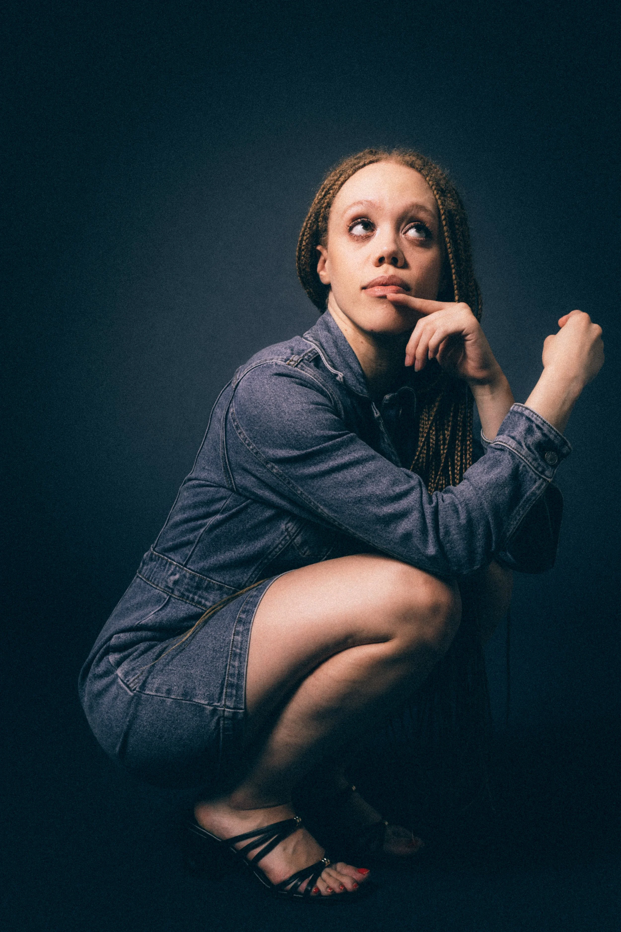 A woman with braided hair is sitting on the ground, wearing a denim jacket and shorts, with black strappy heels and painted red toenails, against a dark background.
