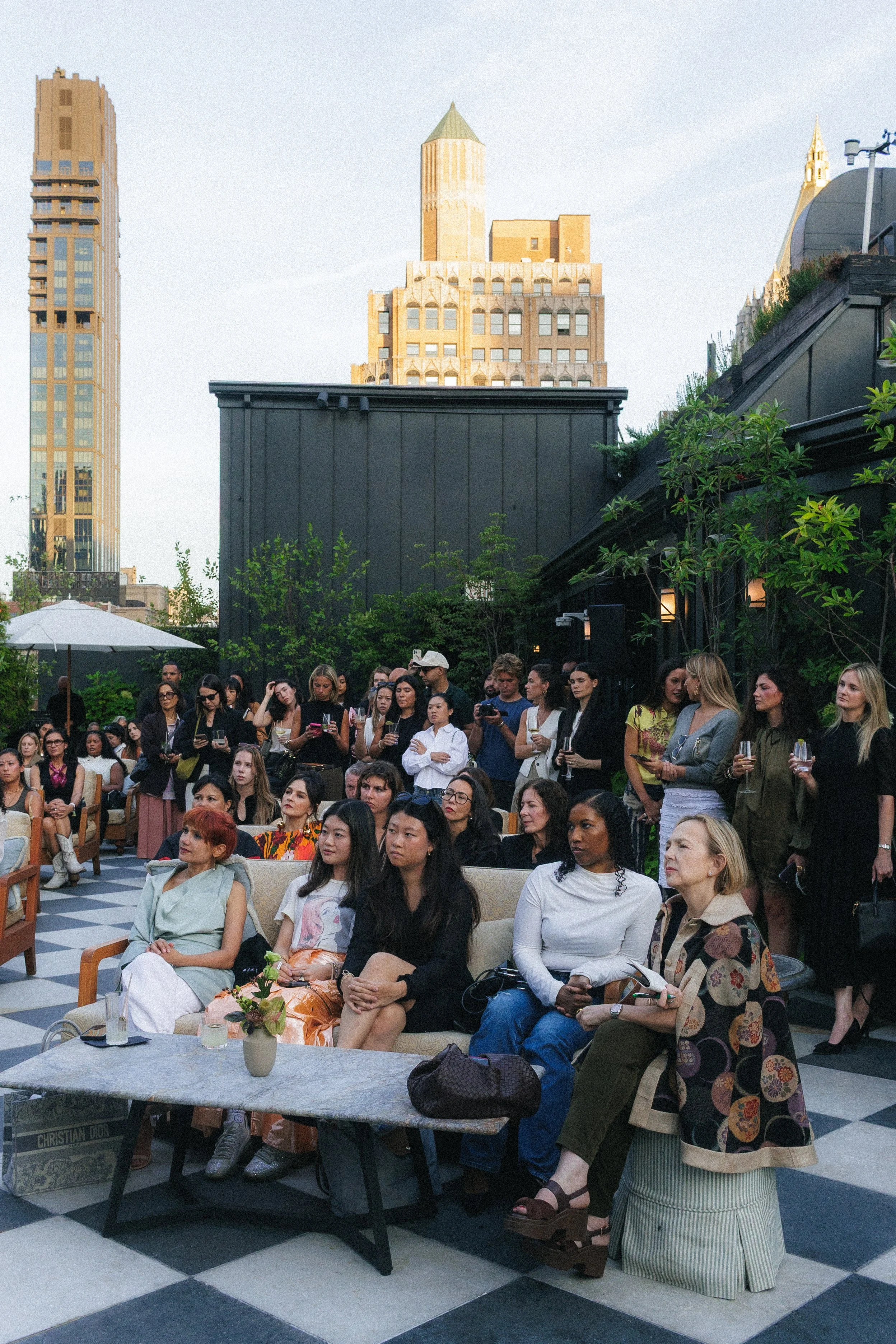 Group of people gathered outdoors on a terrace, seated on chairs and standing, with a city skyline featuring tall buildings in the background.