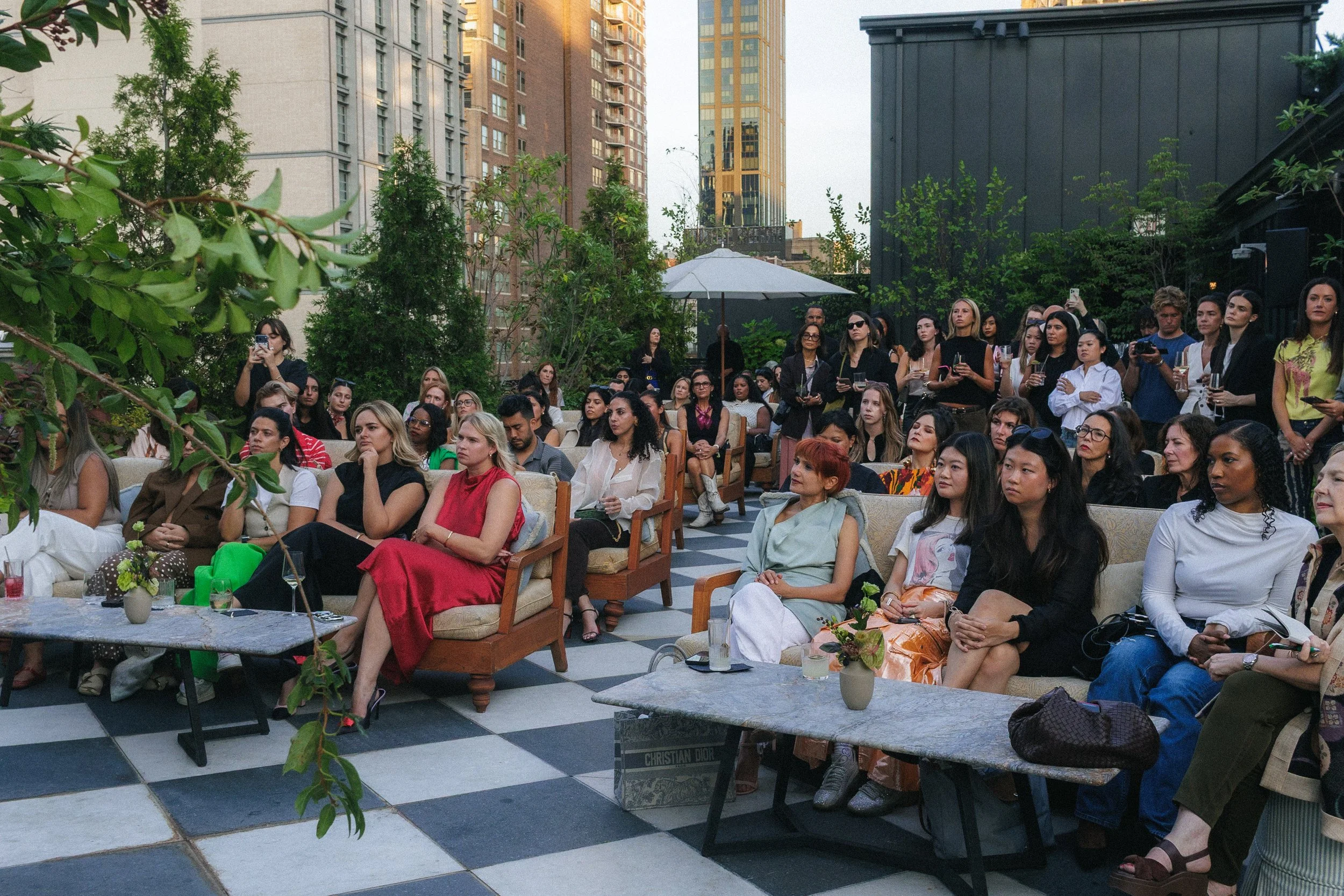 A diverse group of people attending an outdoor event on a rooftop terrace surrounded by tall buildings and greenery, some seated and some standing, with drinks and attentive expressions.