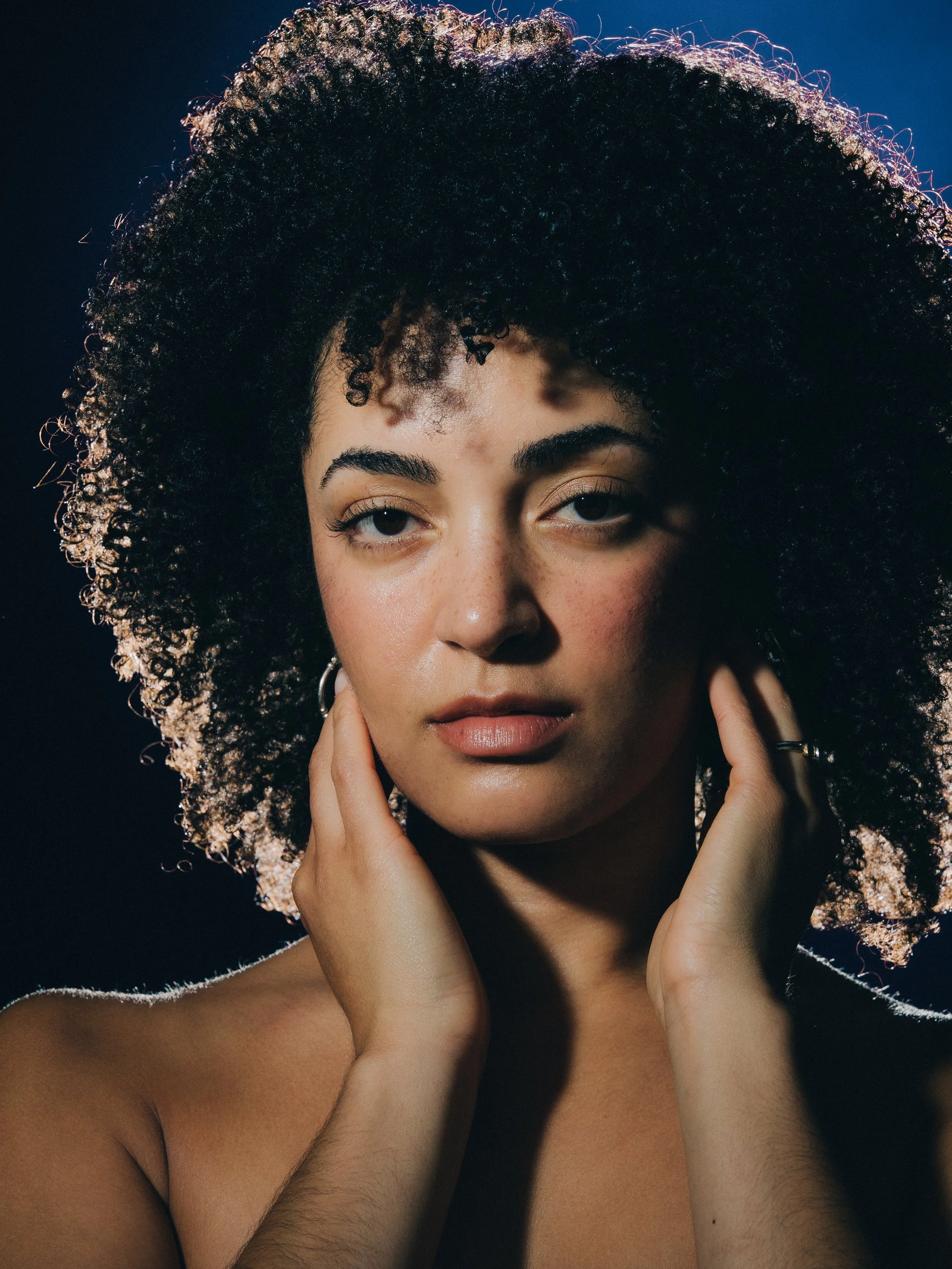 A portrait of a woman with natural curly hair, her hands gently touching her face and neck, against a dark background with soft lighting emphasizing her facial features.