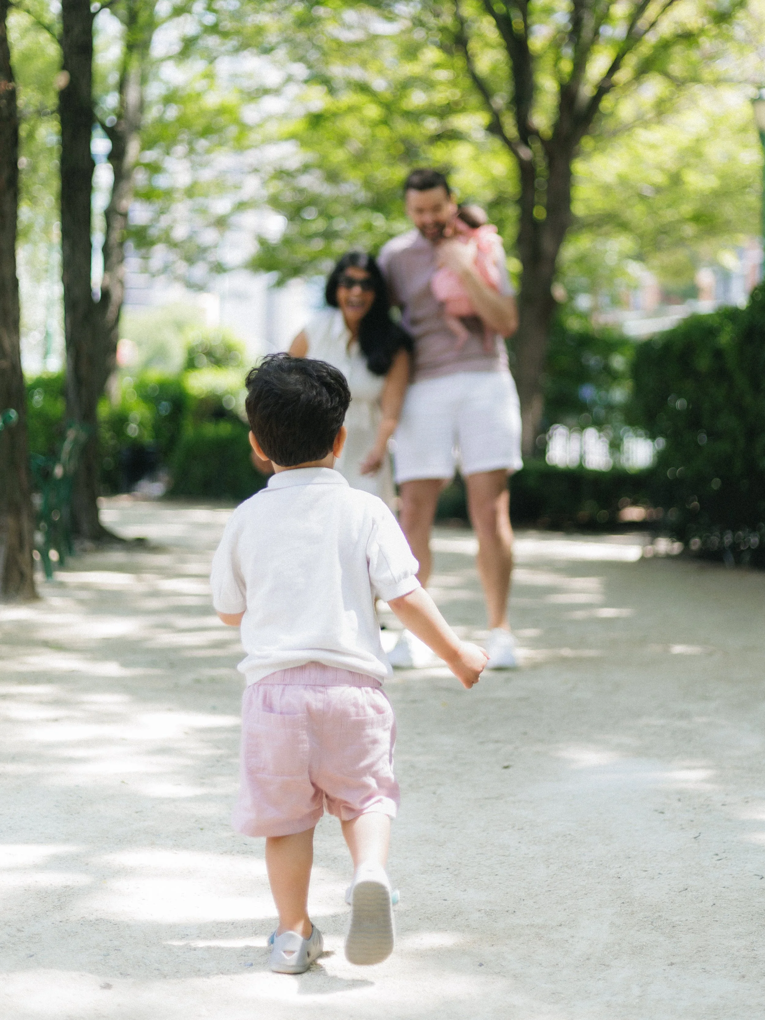 A young child is running on a park pathway with trees and greenery in the background, while a family stands in the distance, smiling.
