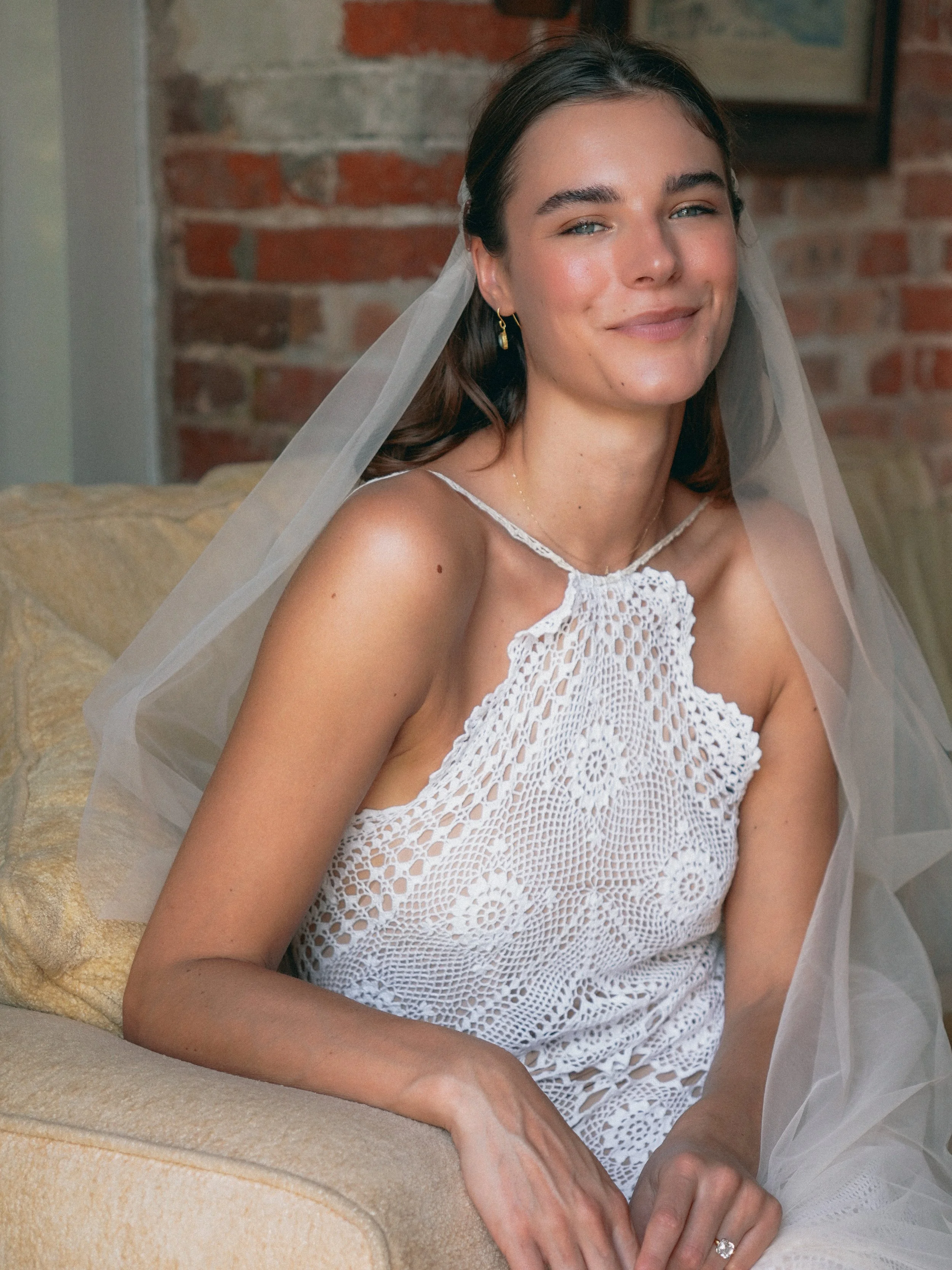 A woman with light skin and dark hair, wearing a white crocheted dress and a delicate necklace, seated on a beige sofa with a brick wall in the background, smiling softly at the camera.