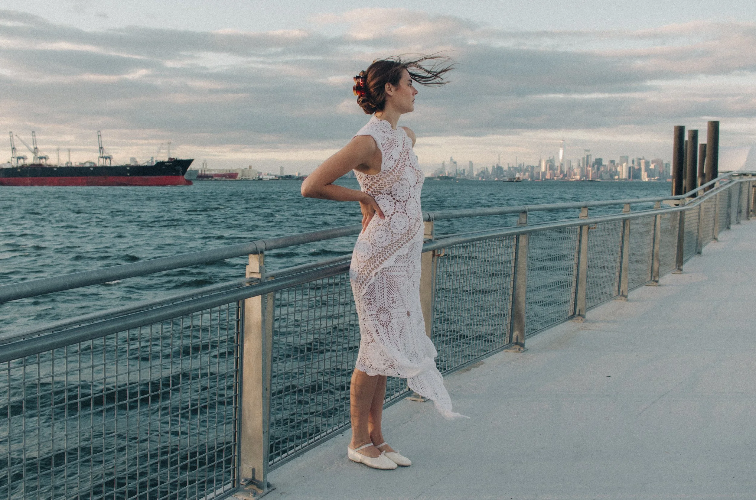 Woman in a white crochet dress and white shoes standing on a pier by the water, with a city skyline and ships in the background, under a cloudy sky.