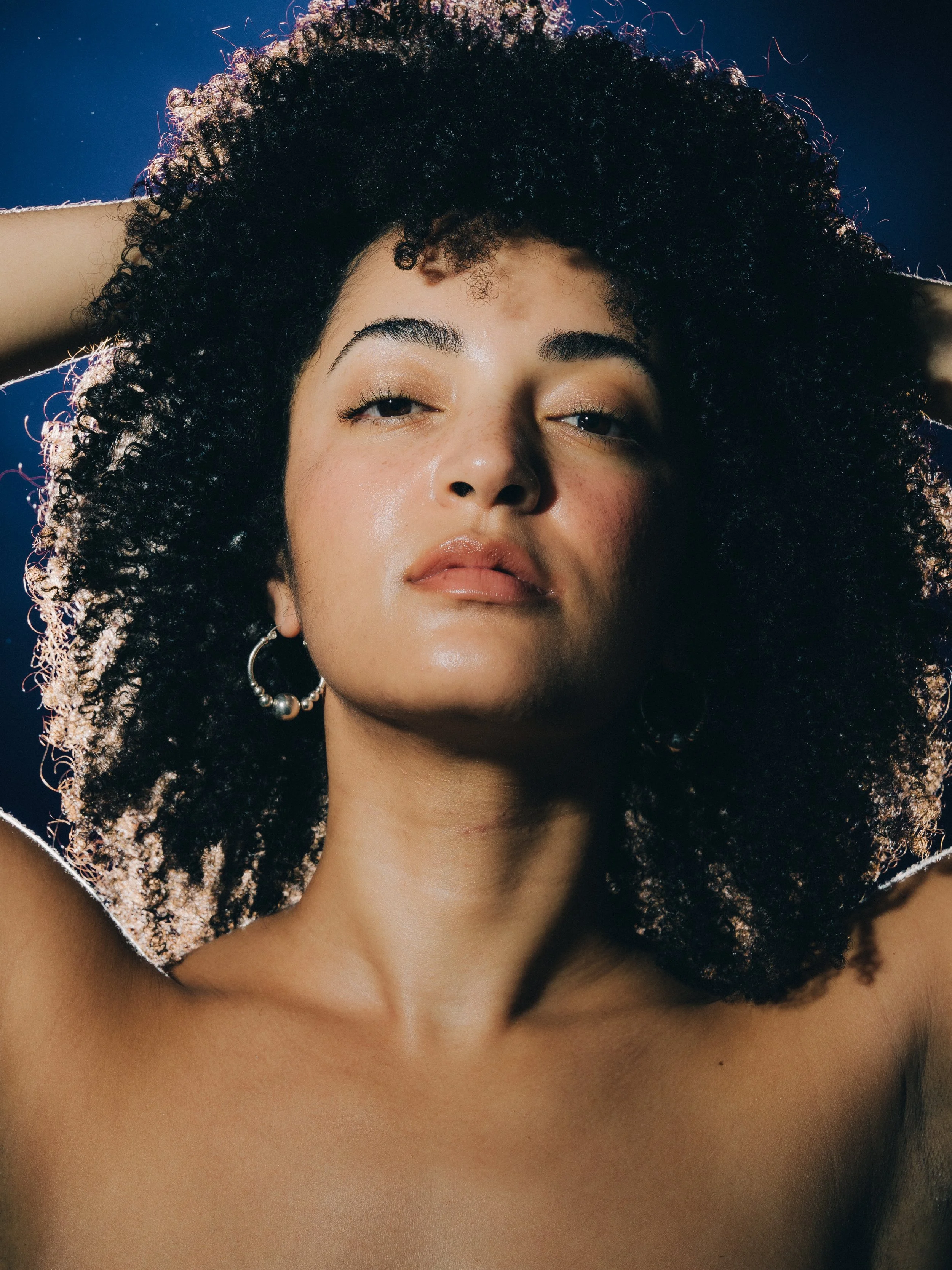 Close-up of a woman with curly black hair, wearing hoop earrings, with a slight smile and radiant skin.