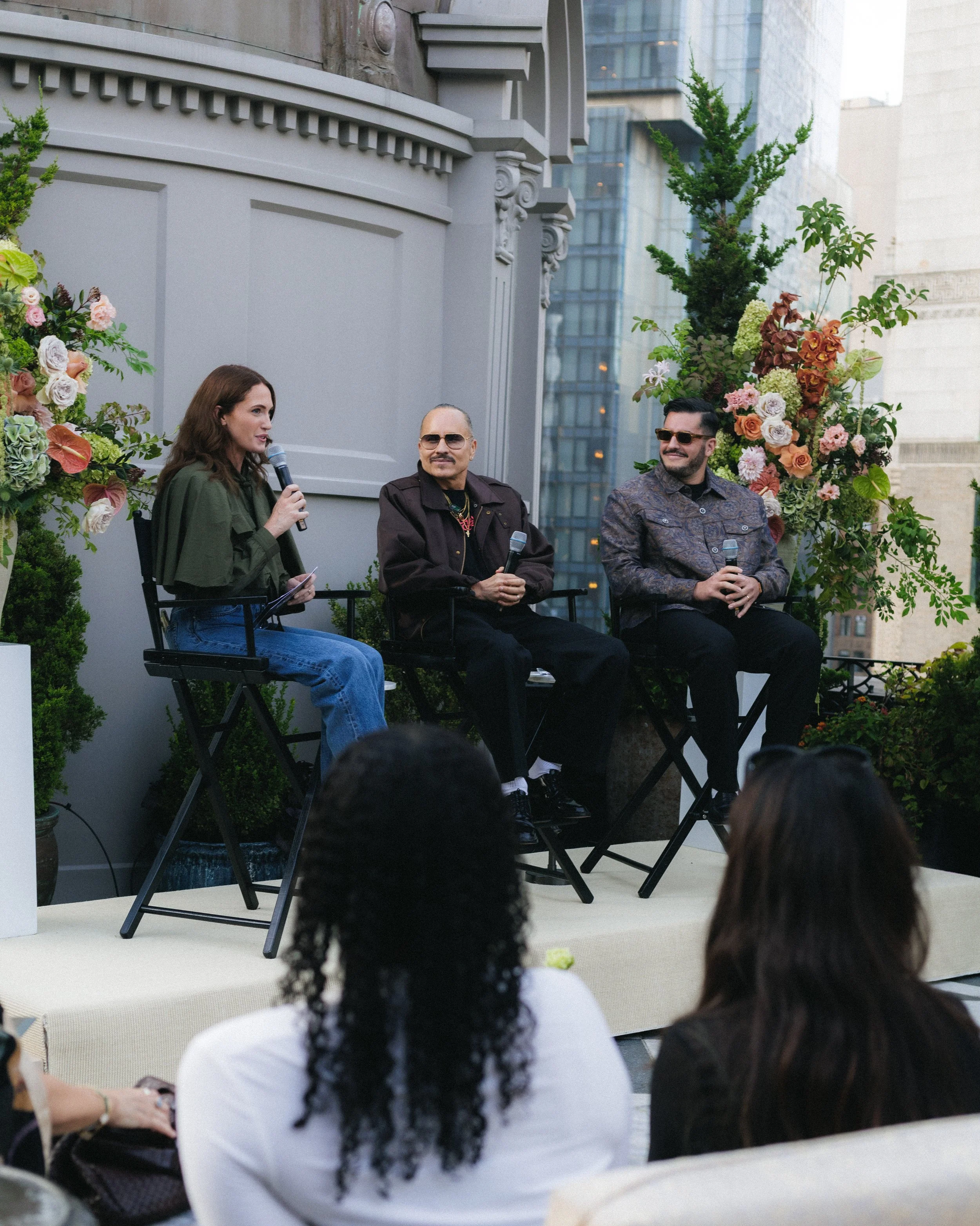 A woman with long, wavy brown hair, wearing a green jacket, is speaking into a microphone at an outdoor event. She is sitting on a black folding chair next to two men, who are also seated and holding microphones. The man in the middle has short, dark