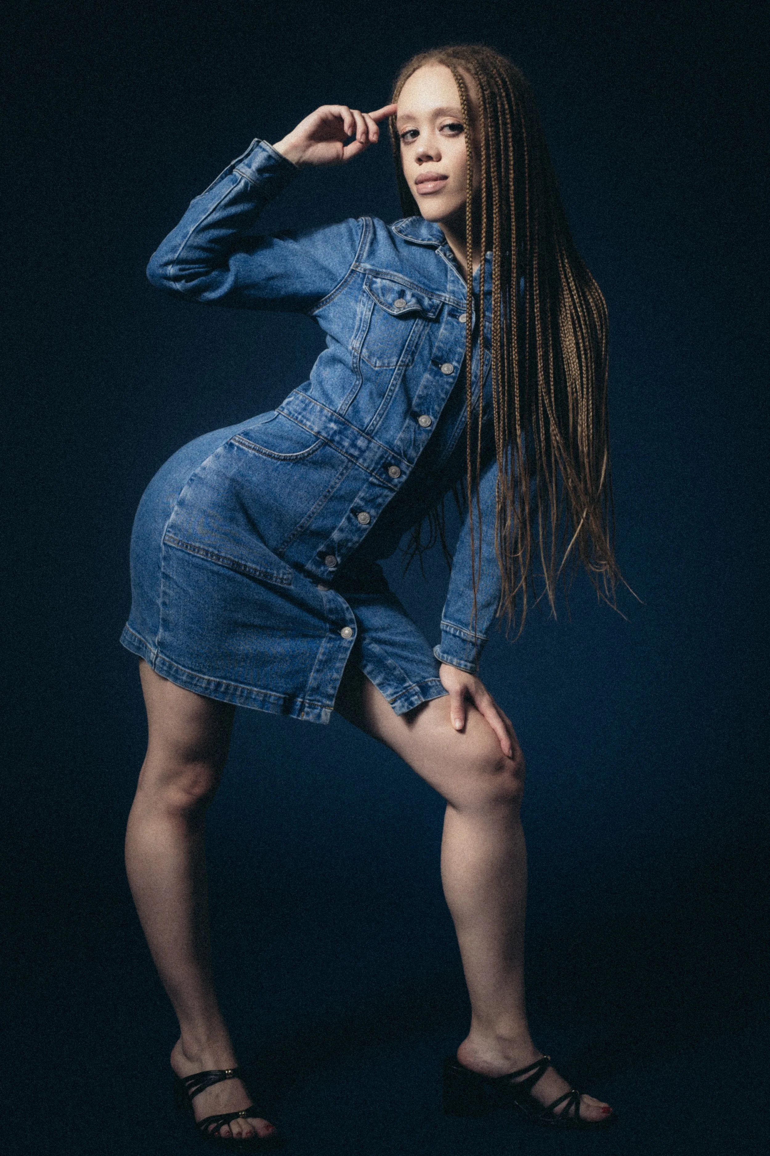 A woman with long braided hair wearing a denim jacket and denim skirt, black high heeled sandals, posing with one hand on her bent knee and the other touching her forehead, against a dark background.