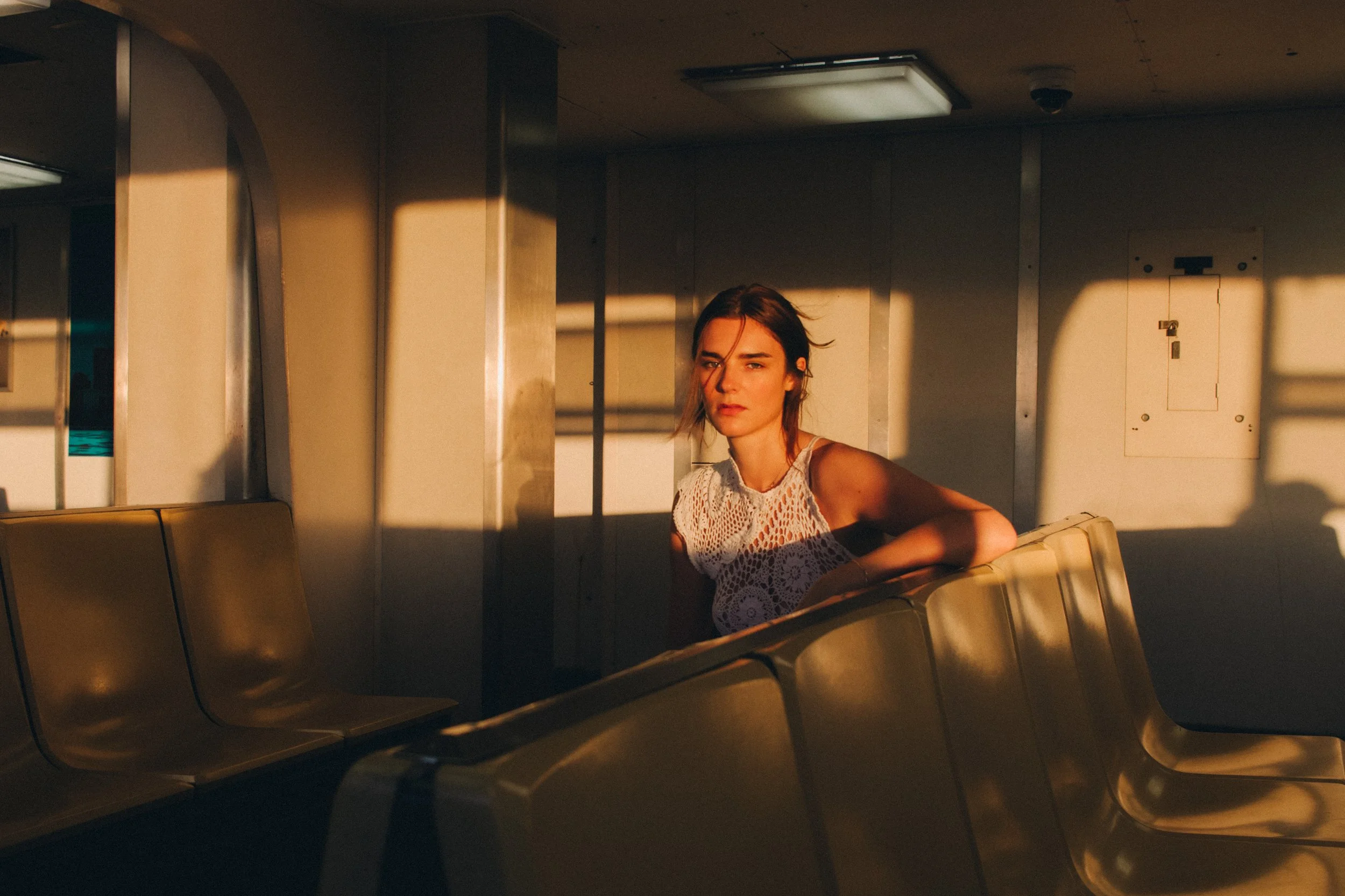 A young woman with dark hair and fair skin sitting on a beige bench in an empty indoor space, illuminated by warm sunlight through a window, creating shadows on the wall behind her.