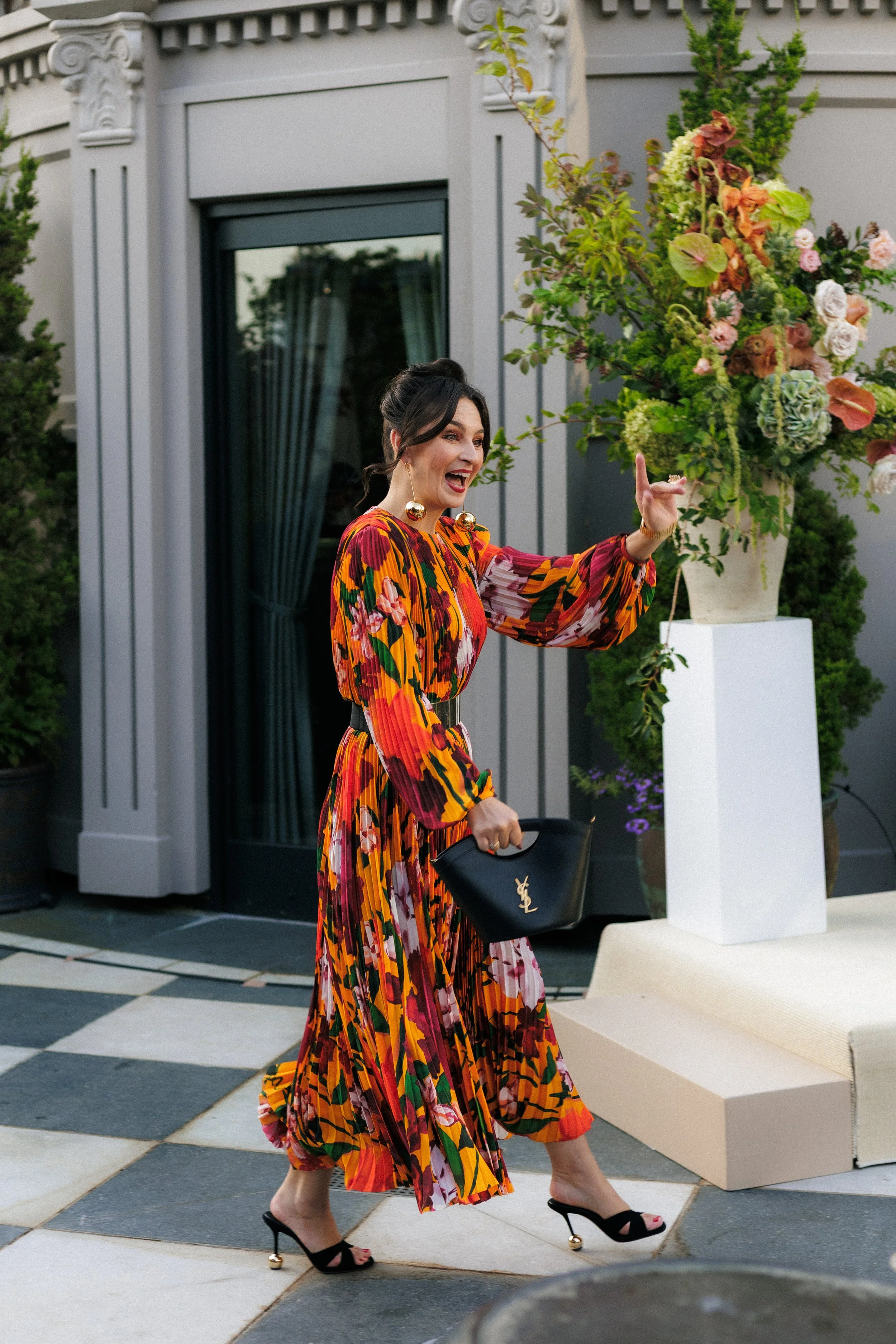 A woman with dark hair styled in an updo, wearing a colorful, floral-printed long dress and black high heels with gold embellishments, smiling and gesturing with her hand while carrying a black Saint Laurent handbag. She is outdoors near a building w