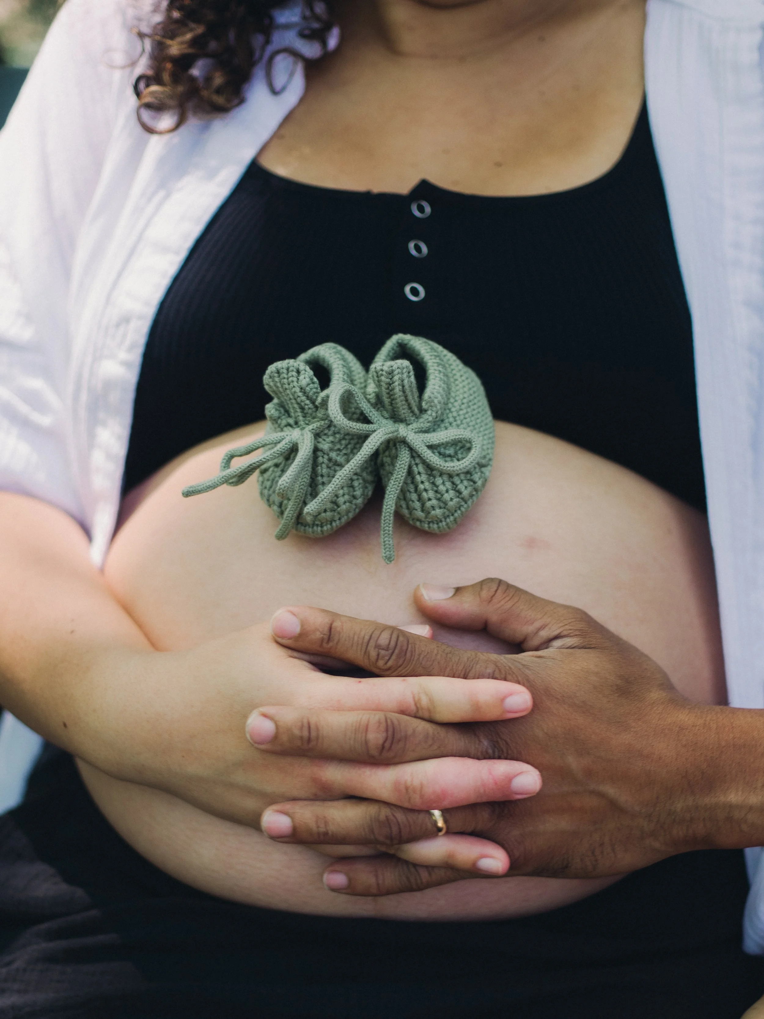A woman is holding her pregnant belly with both hands, with her shirt slightly lifted to reveal her stomach, and a small pair of knitted baby booties resting on her belly.