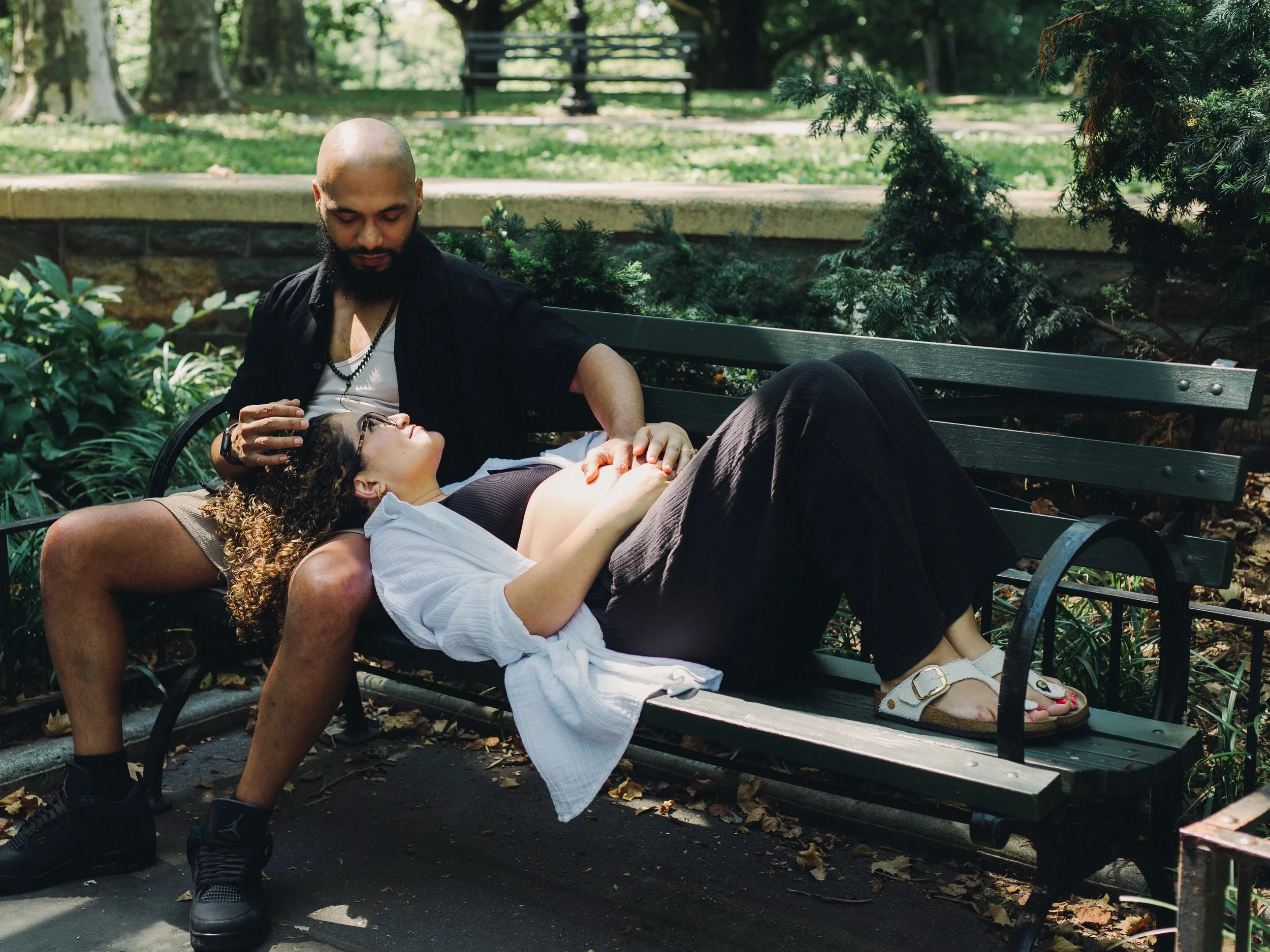 A man with a bald head and beard, wearing a black shirt and black pants, sits on a park bench with a woman with curly hair, glasses, and light skin, lying on his lap. The woman is wearing a white dress and sandals. They are in a green park with trees