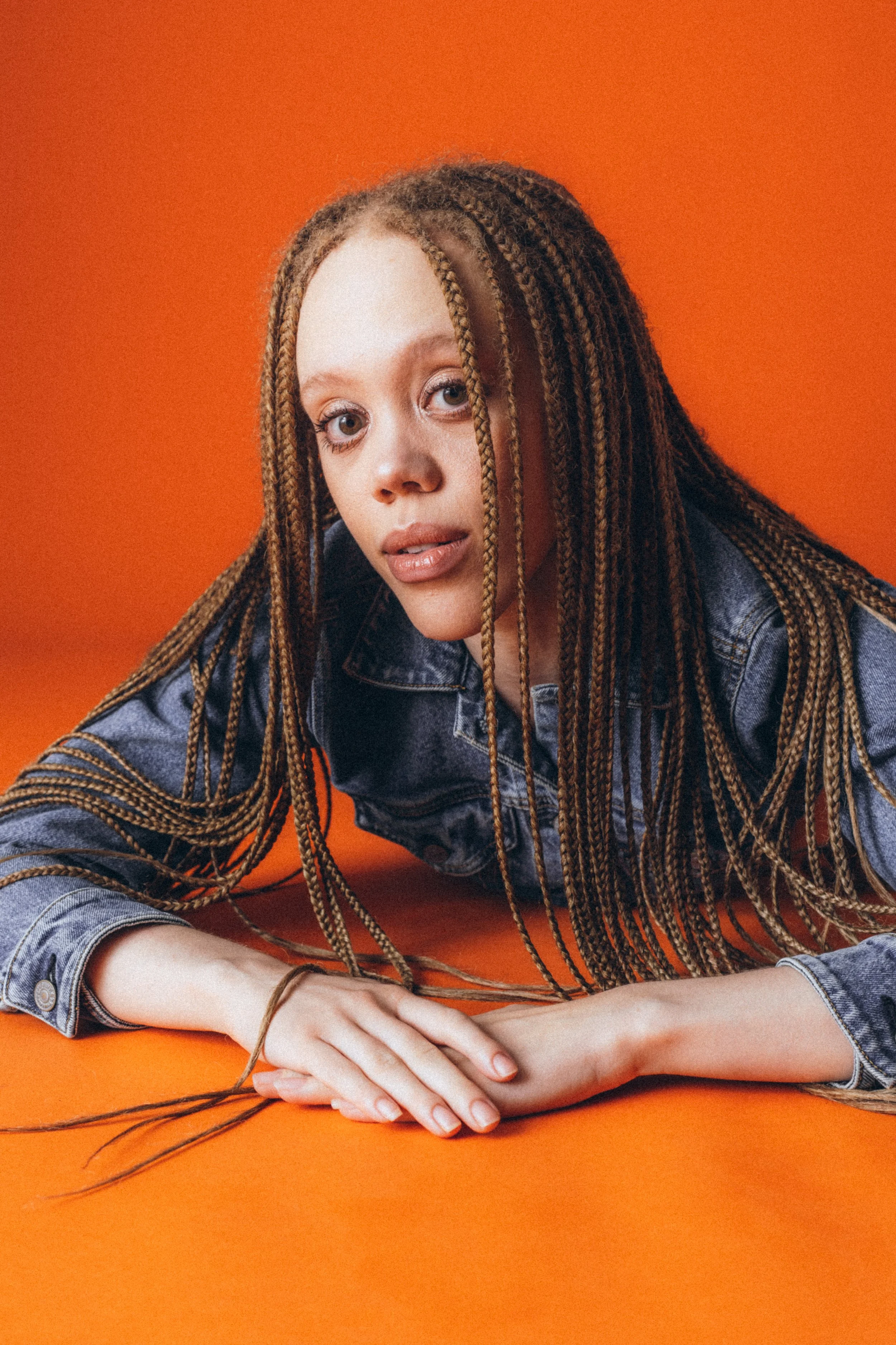 A young woman with braided hair, wearing a denim jacket, lying on an orange surface, with a matching orange background, looking into the camera.