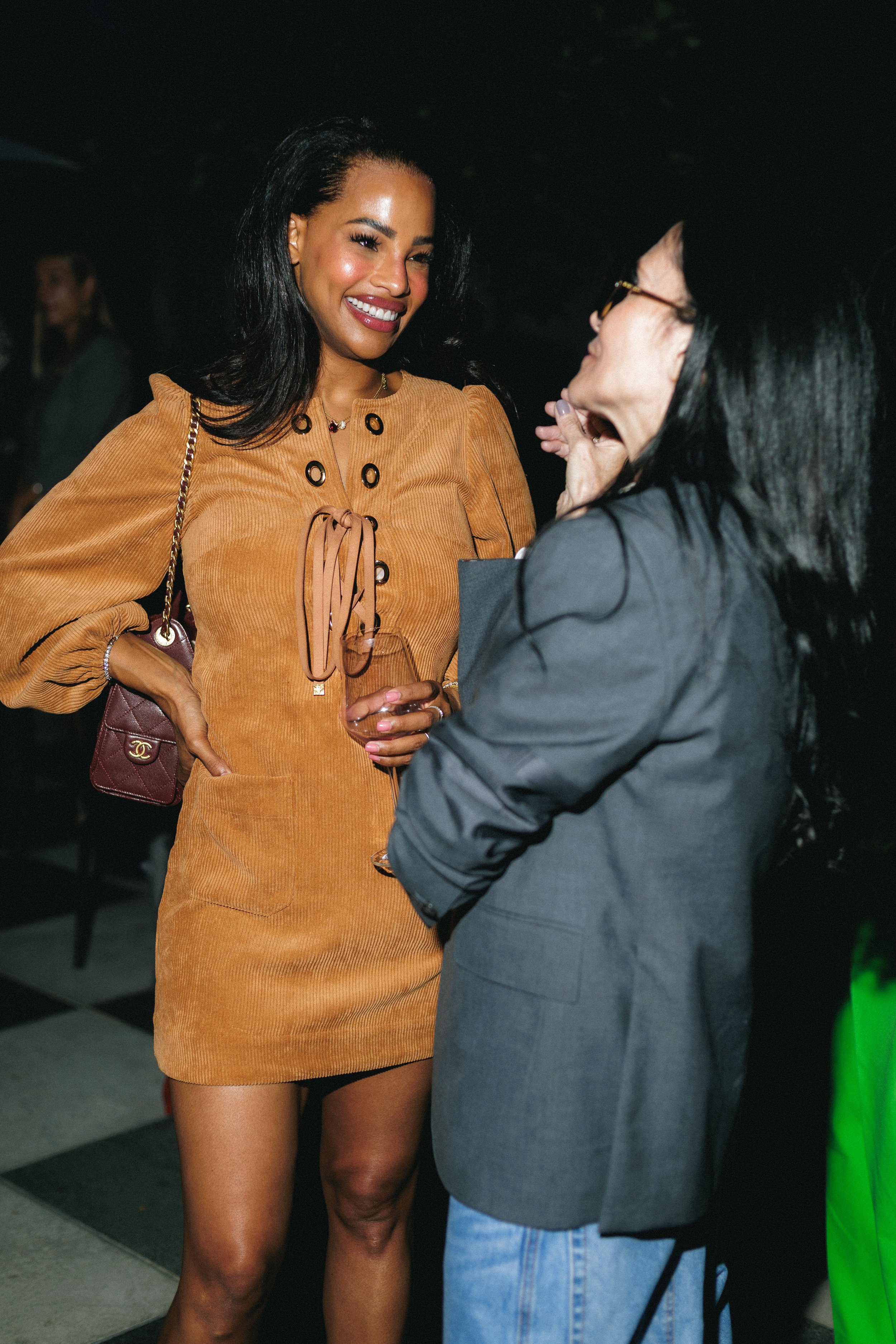 Two women are engaging in conversation at a social event. The woman on the left is smiling, wearing a tan dress with black and gold collar details, holding a glass of drink, and carrying a Chanel purse. The woman on the right, seen from the side, is 