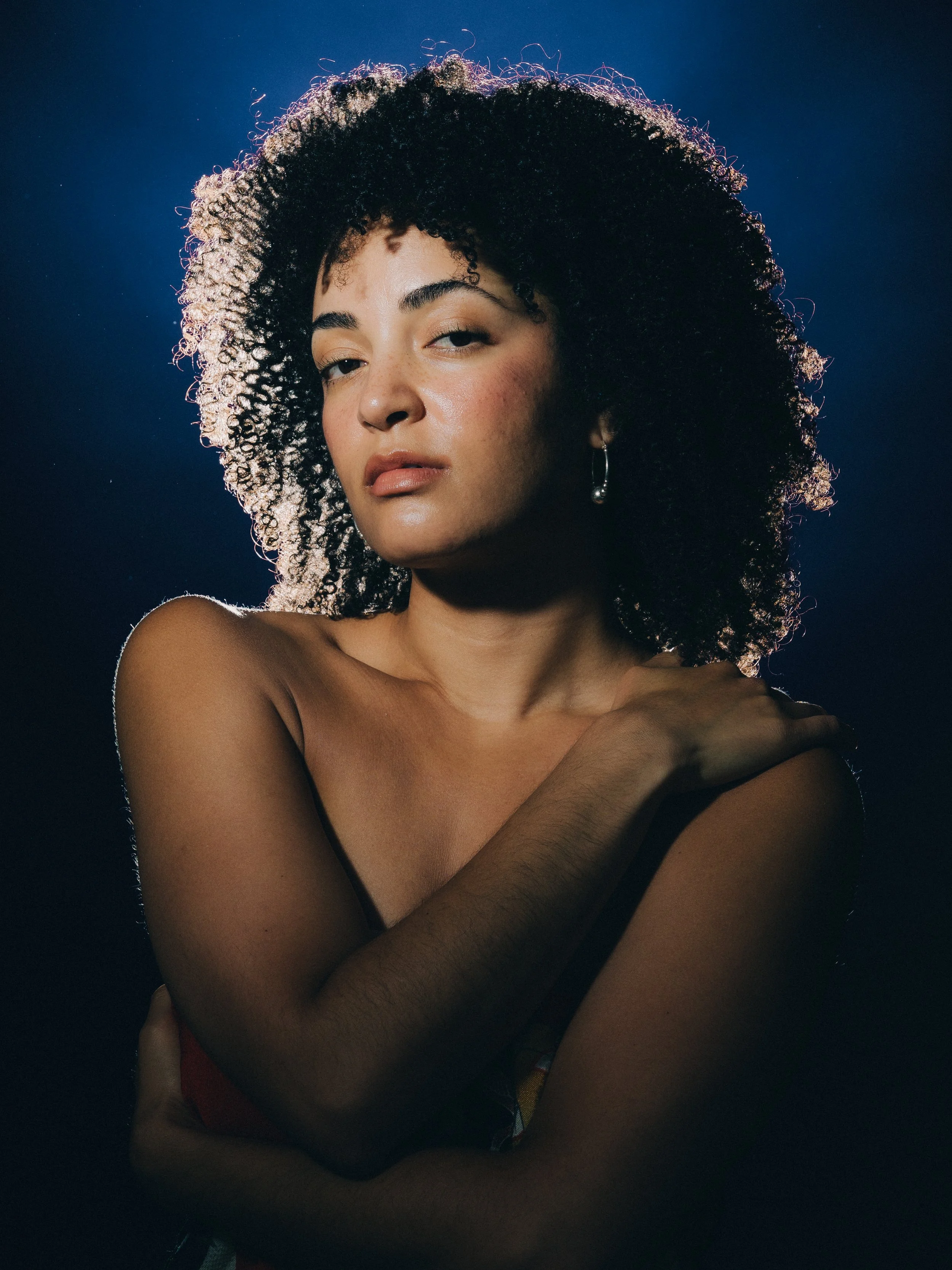 Portrait of a woman with curly dark hair, wearing pearl earrings, with a dark background, looking at the camera with a neutral expression.
