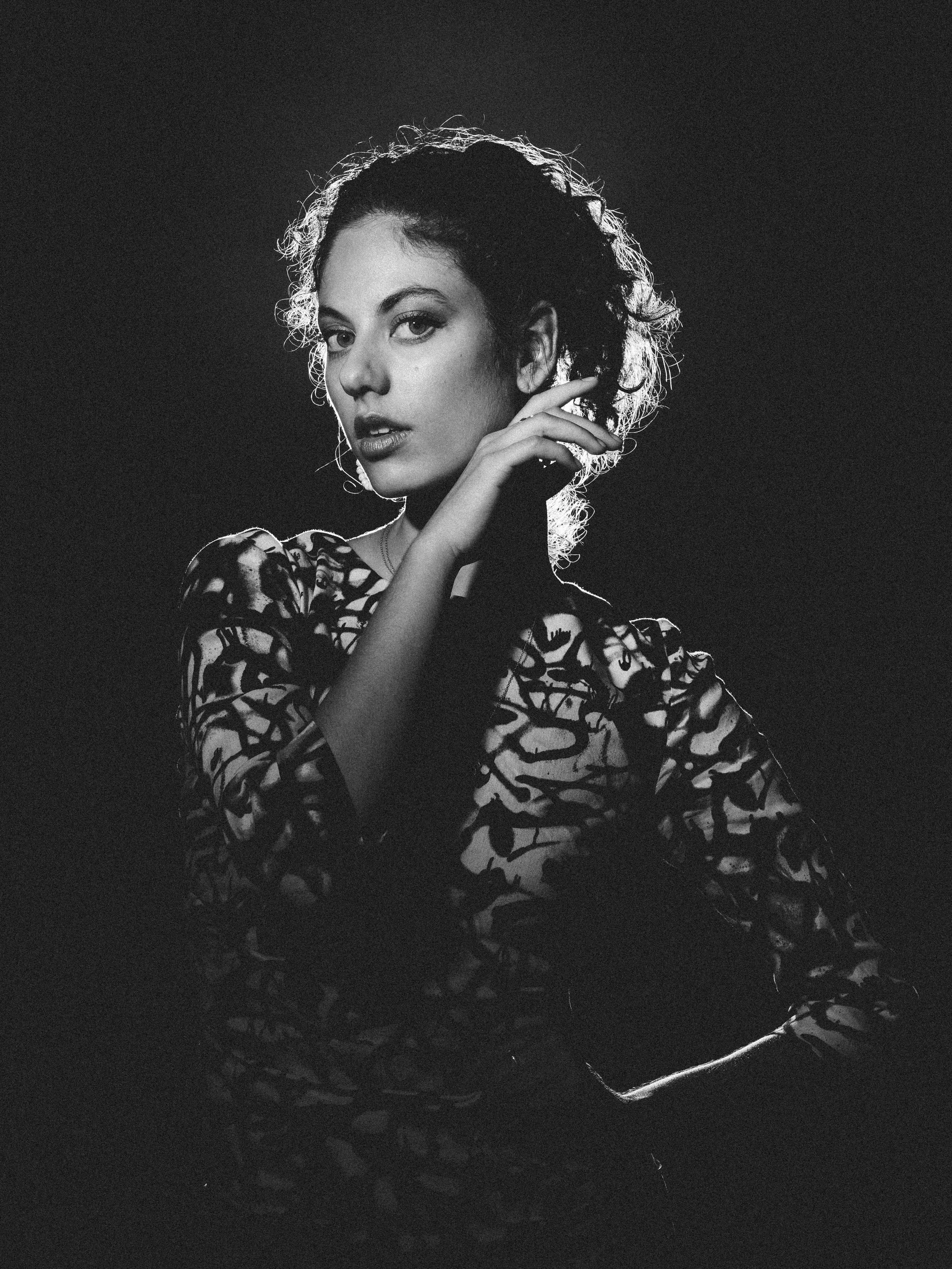 Black and white portrait of a woman with short curly hair, wearing a patterned top, with one hand touching her face and looking at the camera.