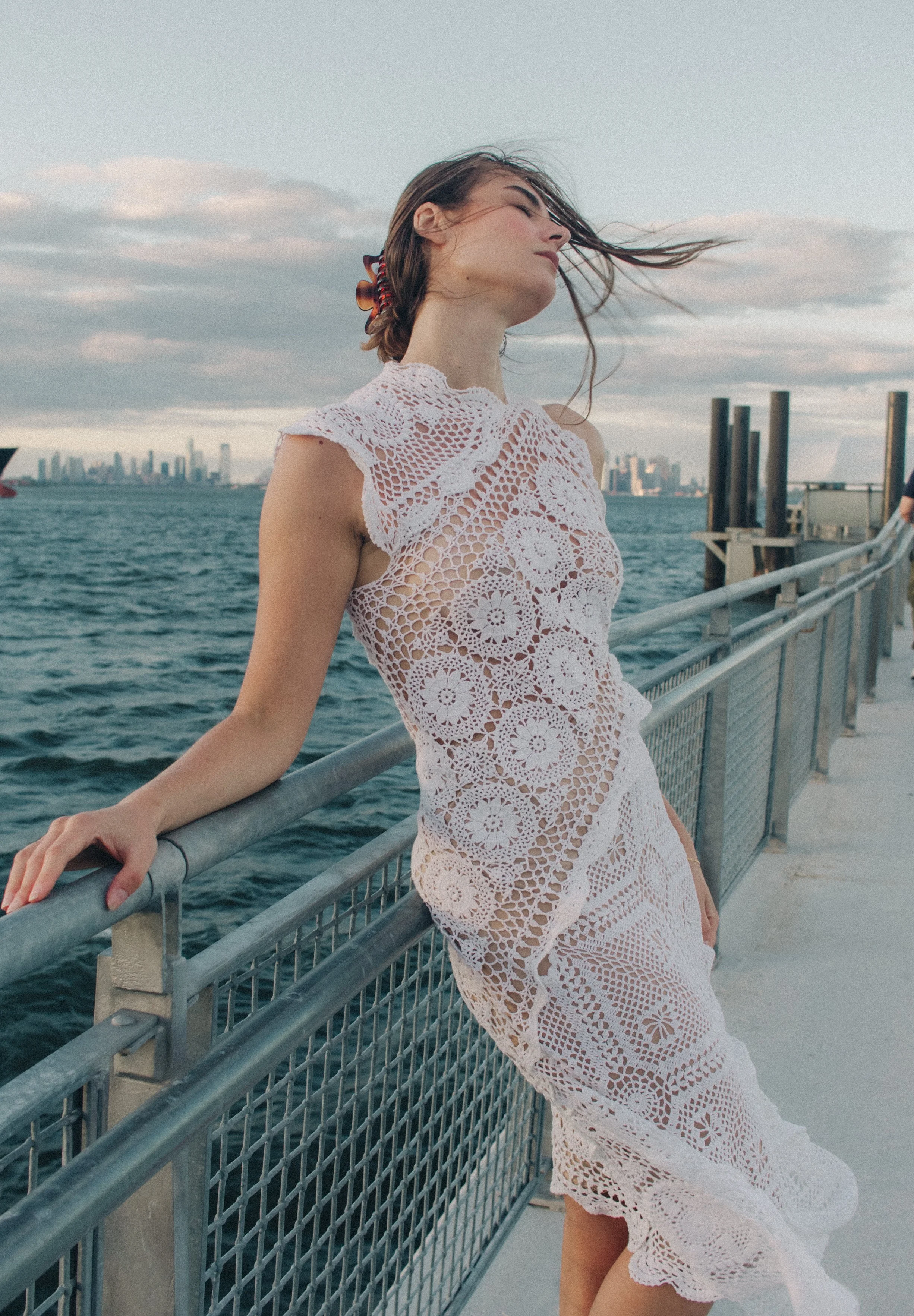 A woman with long hair tied with a hairband, wearing a white lace dress, stands on a pier by the water with a city skyline in the background. She has her head tilted back and her eyes closed, with wind blowing her hair.