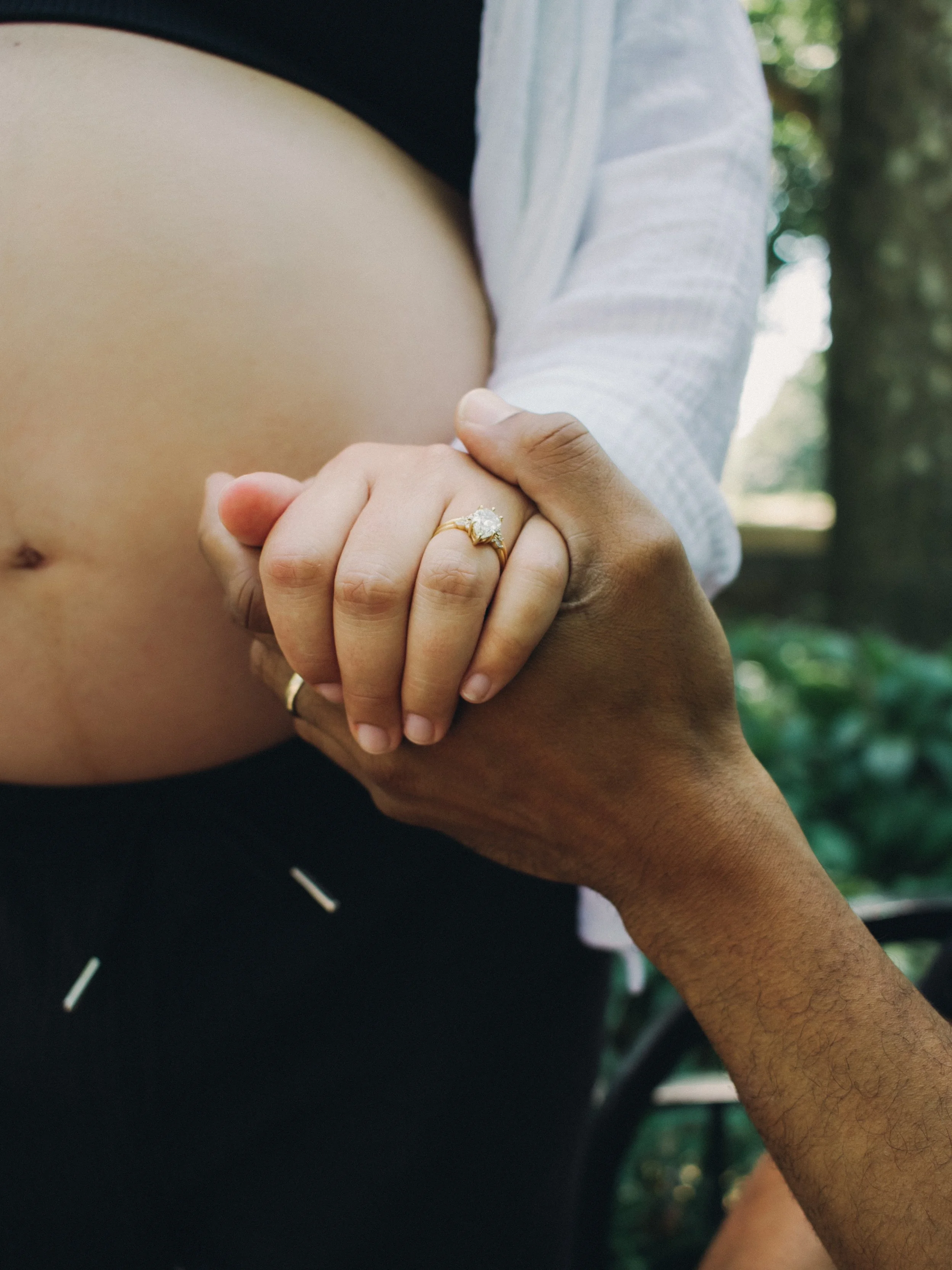 Close-up of two people's hands, one with a diamond engagement ring, holding a pregnant belly outdoors with trees in the background.