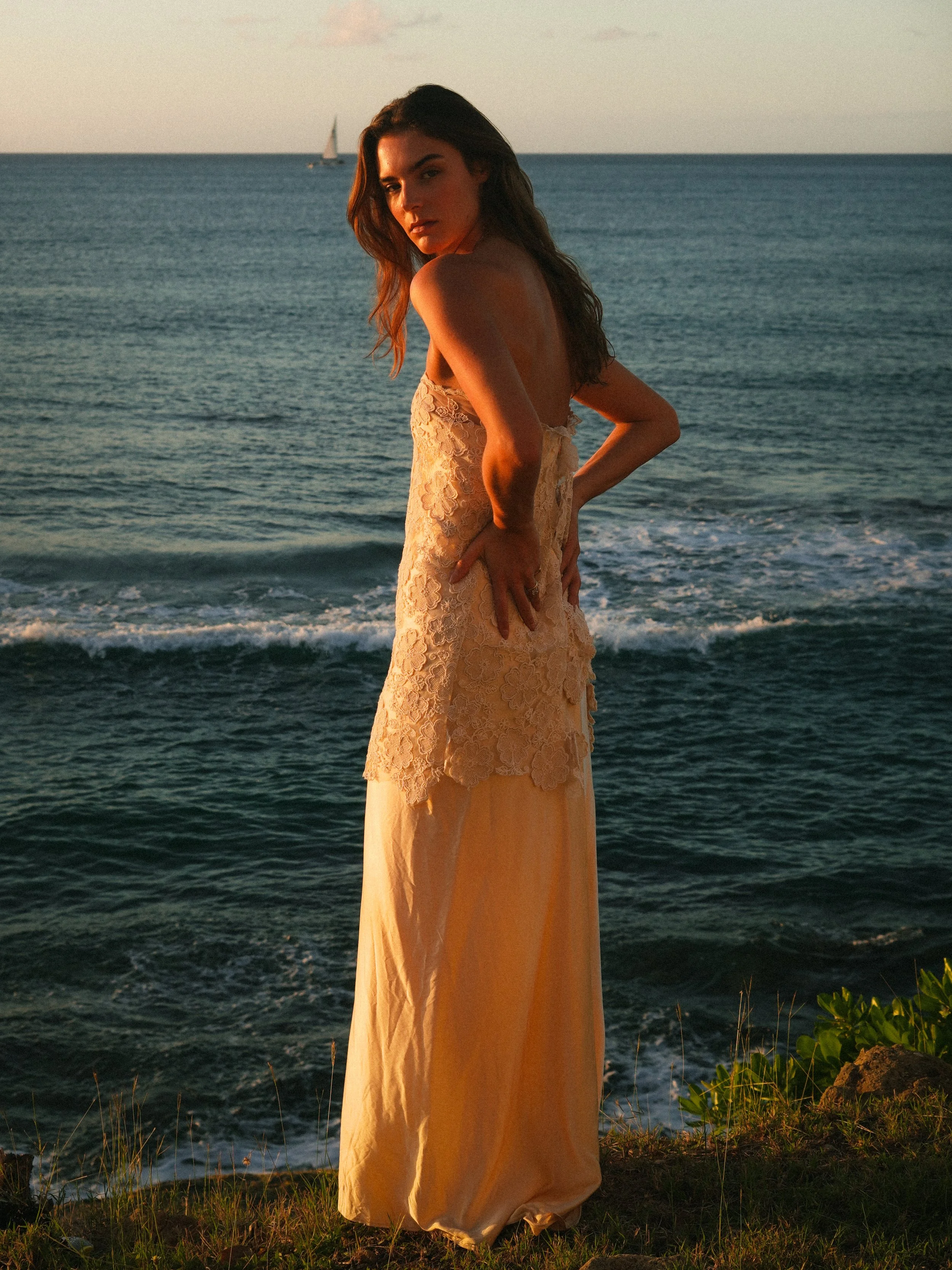 A woman in a beige lace dress standing on a grassy edge by the ocean during sunset, with a sailboat in the background.
