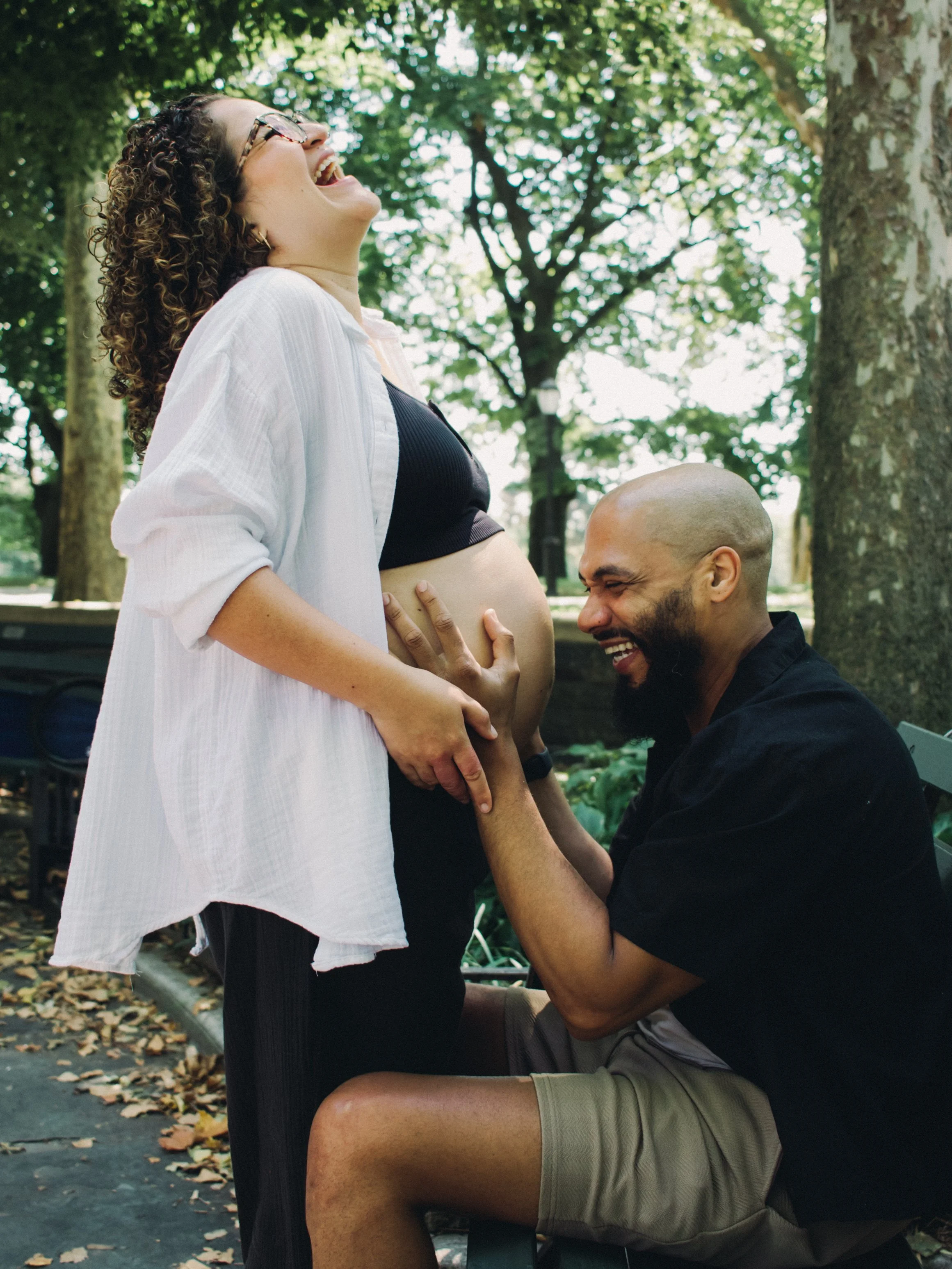 A pregnant woman standing outdoors, smiling and laughing, with a man kneeling before her, touching her belly and smiling. They are surrounded by trees and fallen leaves.