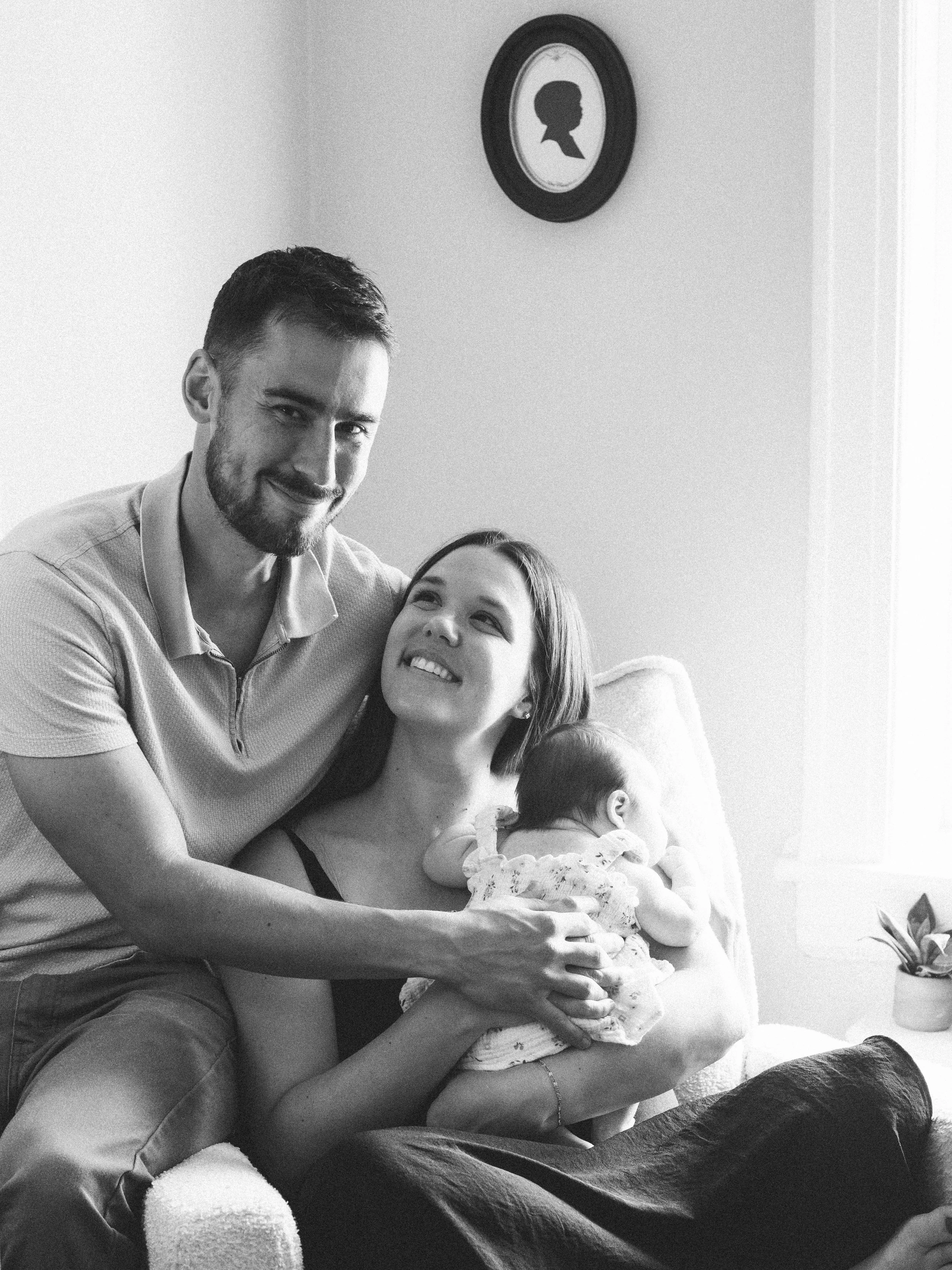 A man and woman with a baby sitting on a couch in a living room, smiling and looking at each other. There is a framed silhouette profile picture on the wall behind them.