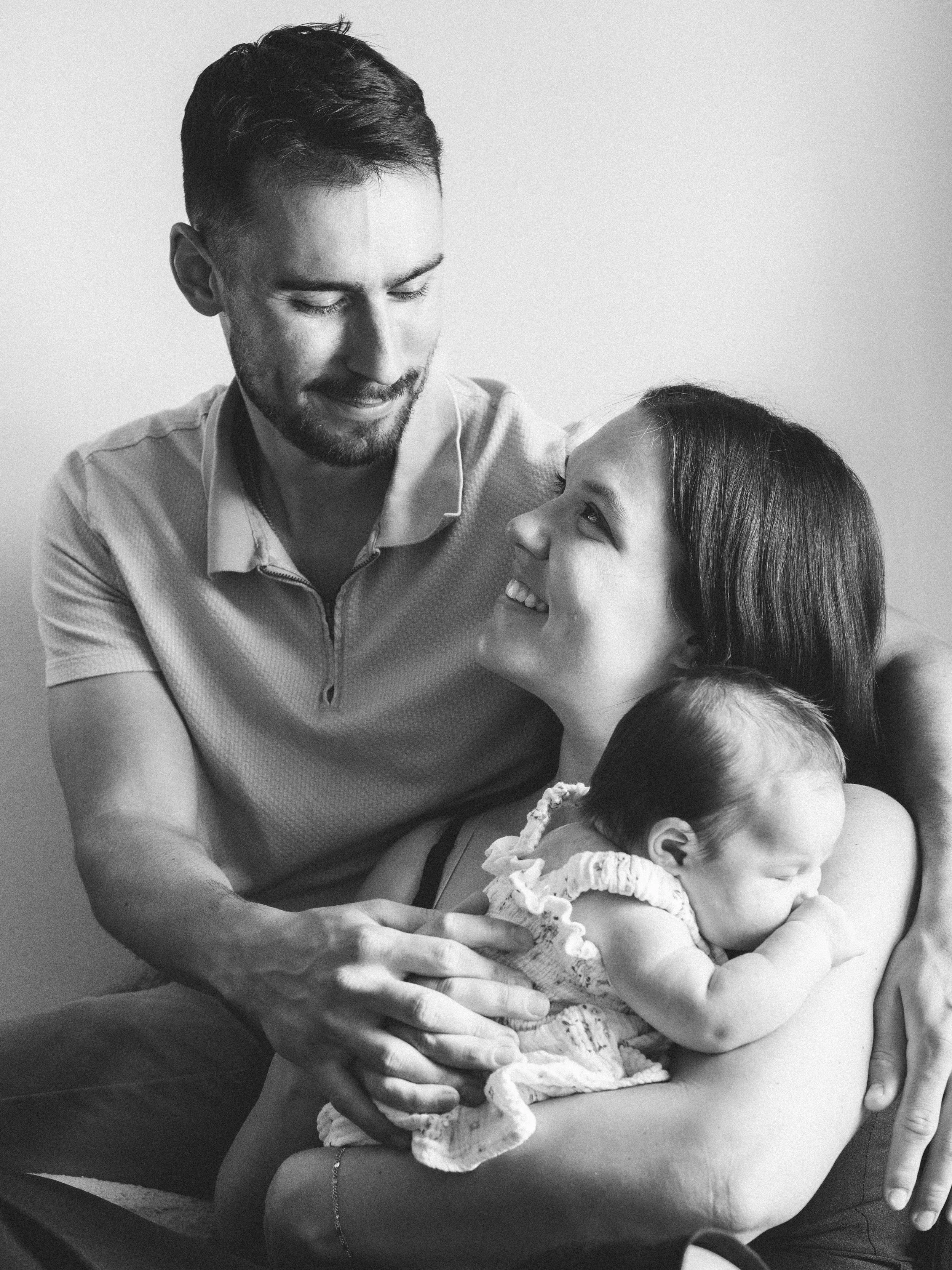 A man with a beard and a woman with long hair are sitting close when the man is smiling and the woman is looking up at him happily. There is a young child, possibly a baby, resting on the woman's lap.