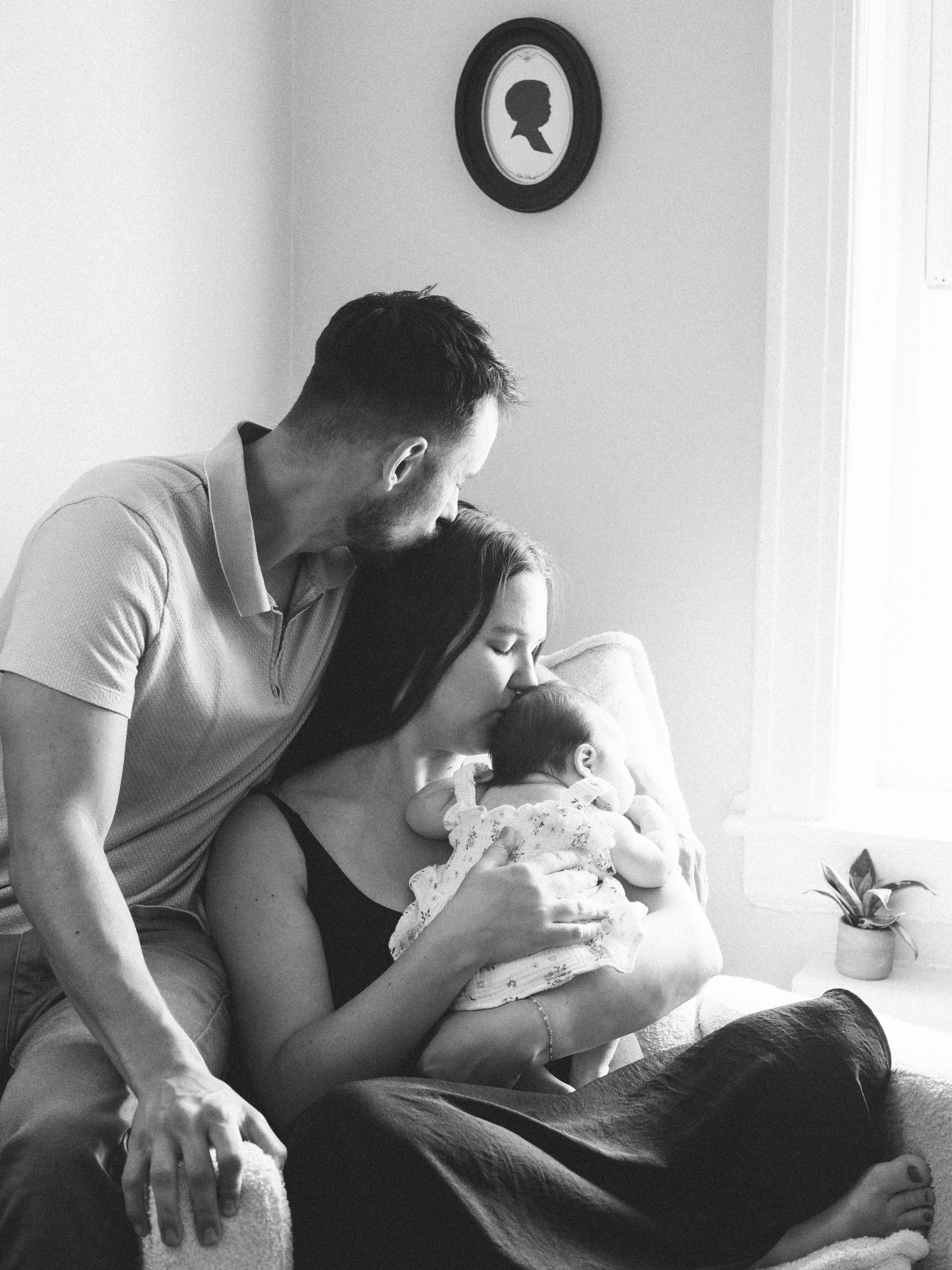 A man and woman sitting on a couch with a baby. The man is leaning over the woman, who is holding the baby. There is a framed silhouette portrait on the wall and a plant on a nearby table.
