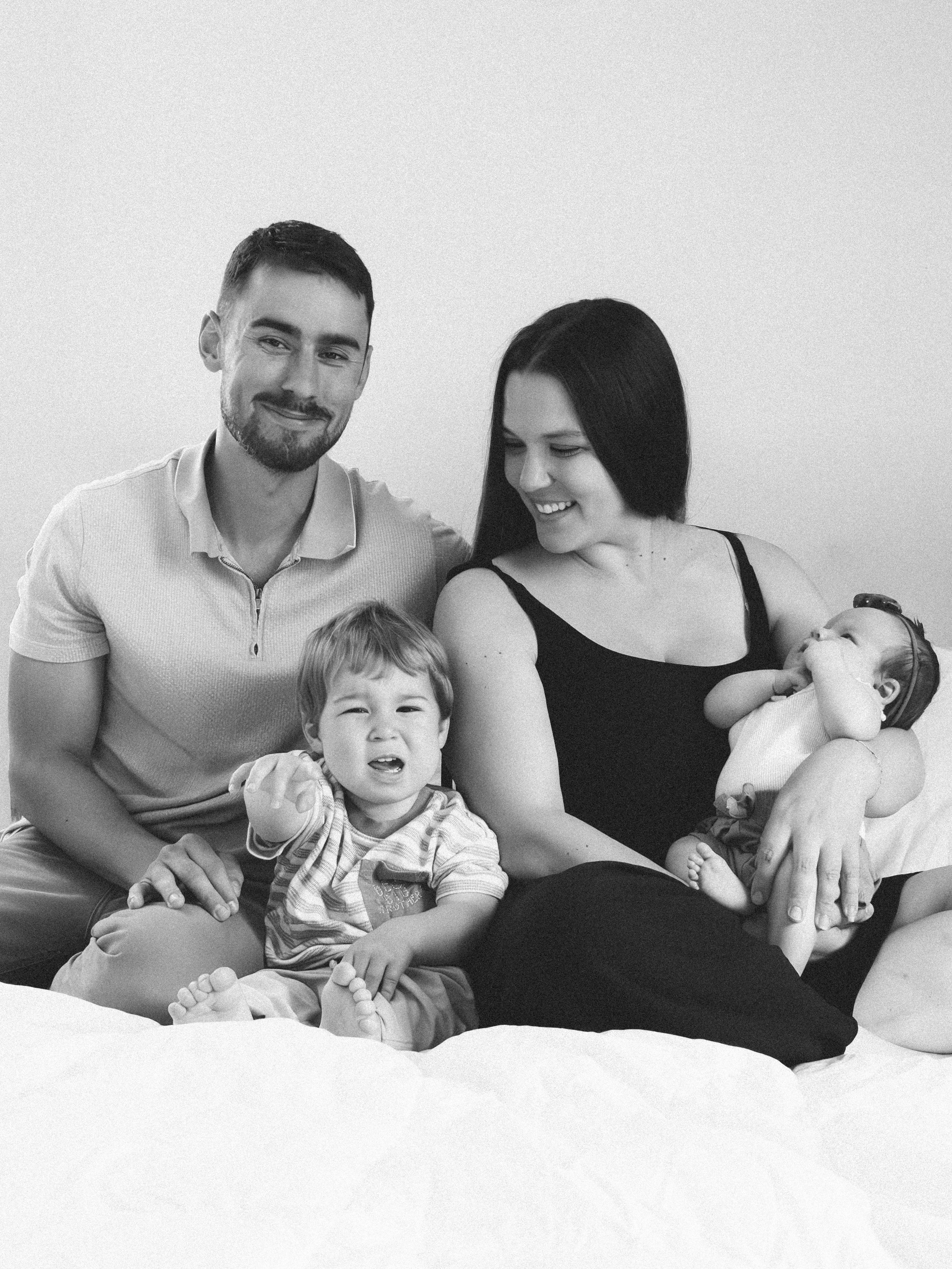 Black and white photo of a smiling man and woman with two young children, all sitting together on a bed.