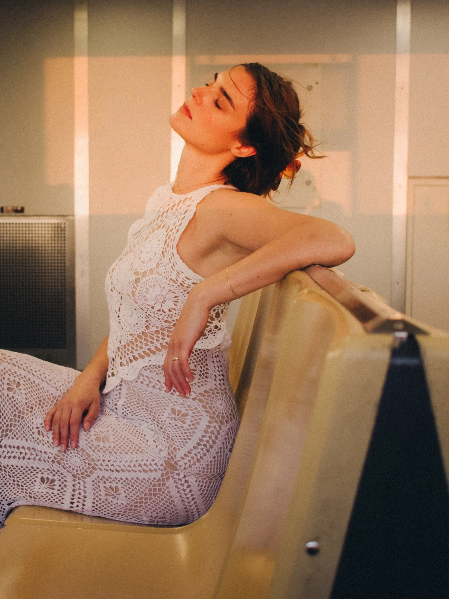 A woman with short brown hair wearing a white crocheted dress sitting on a beige bench with eyes closed, head tilted back, in a warm-lit room.