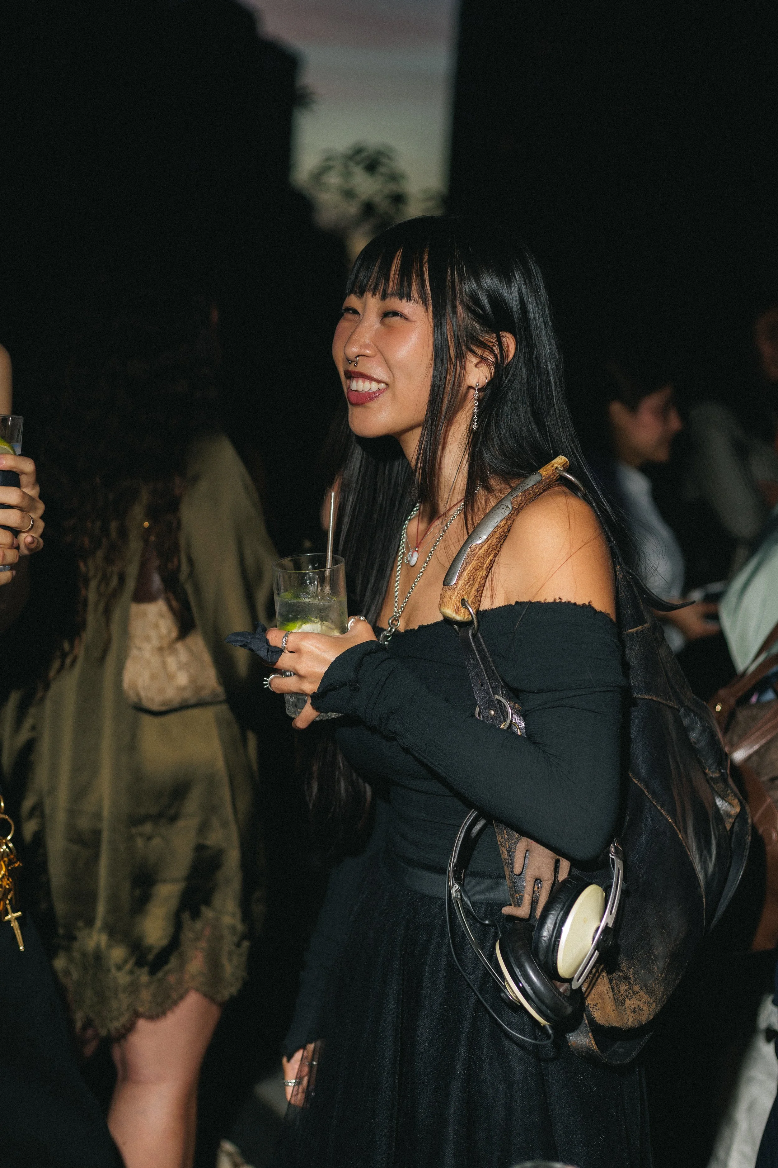 A woman with long black hair with orange streaks, wearing earrings, a septum piercing, and a black off-shoulder top, smiling and holding a drink at a night event, with a background of dark, blurred people and a projection screen.