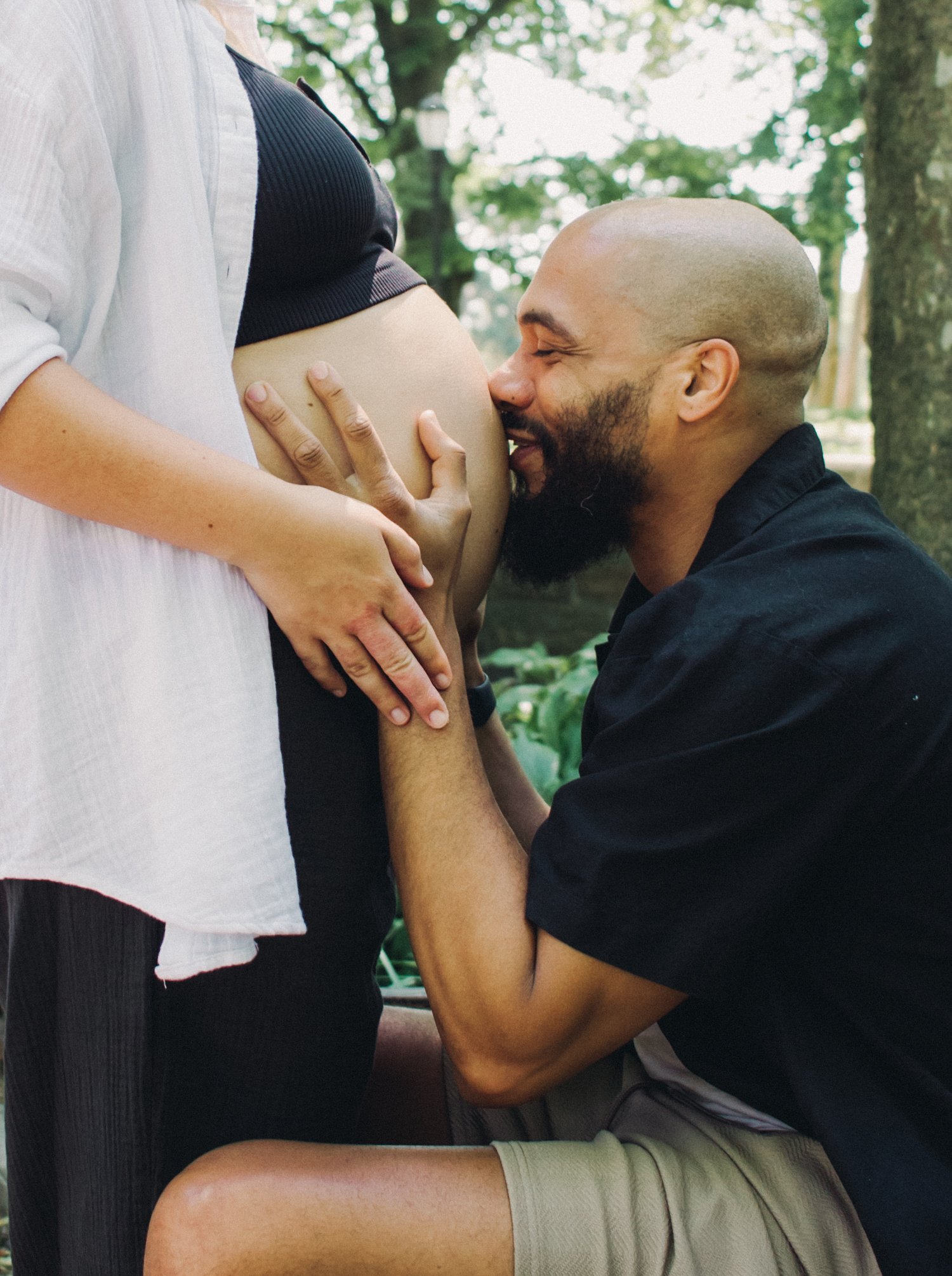 A man kneeling and kissing a pregnant woman’s belly outdoors with trees in the background.