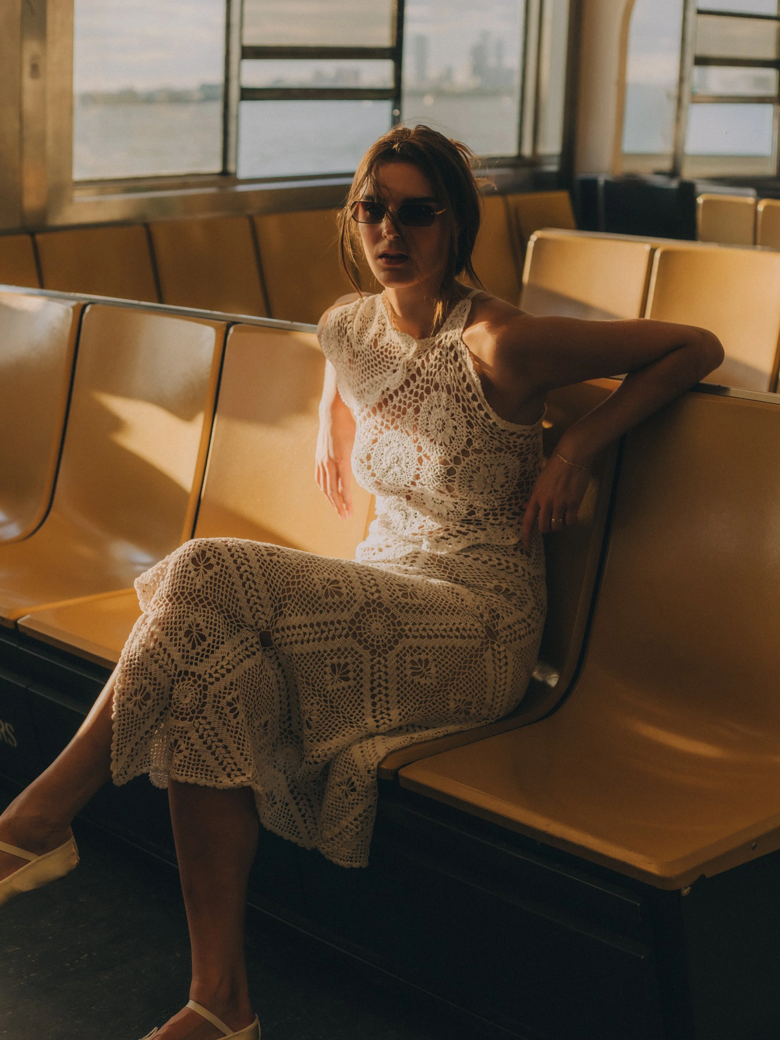 A woman sitting alone on an empty yellow game or waiting area seat in an indoor setting with large windows showing a body of water and city skyline in the background. She is wearing a white crochet dress and sunglasses, with her hair styled in a mess
