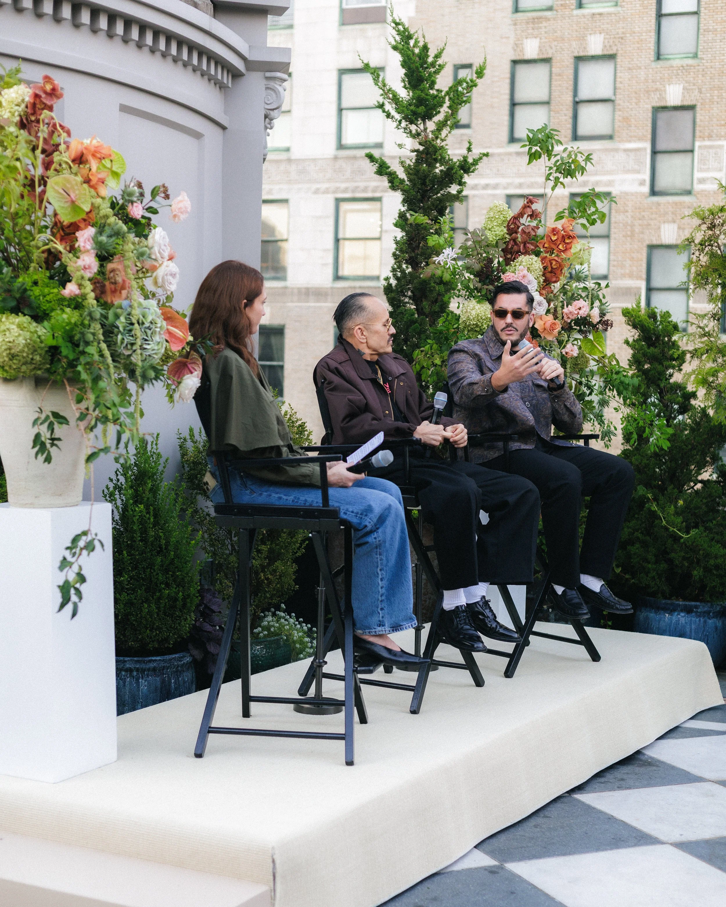 Three people sit on a stage outdoors, engaged in a discussion, with a floral arrangement and greenery behind them. The man on the right wears sunglasses and holds a microphone, gesturing as he speaks. The woman on the left has long brown hair, and th