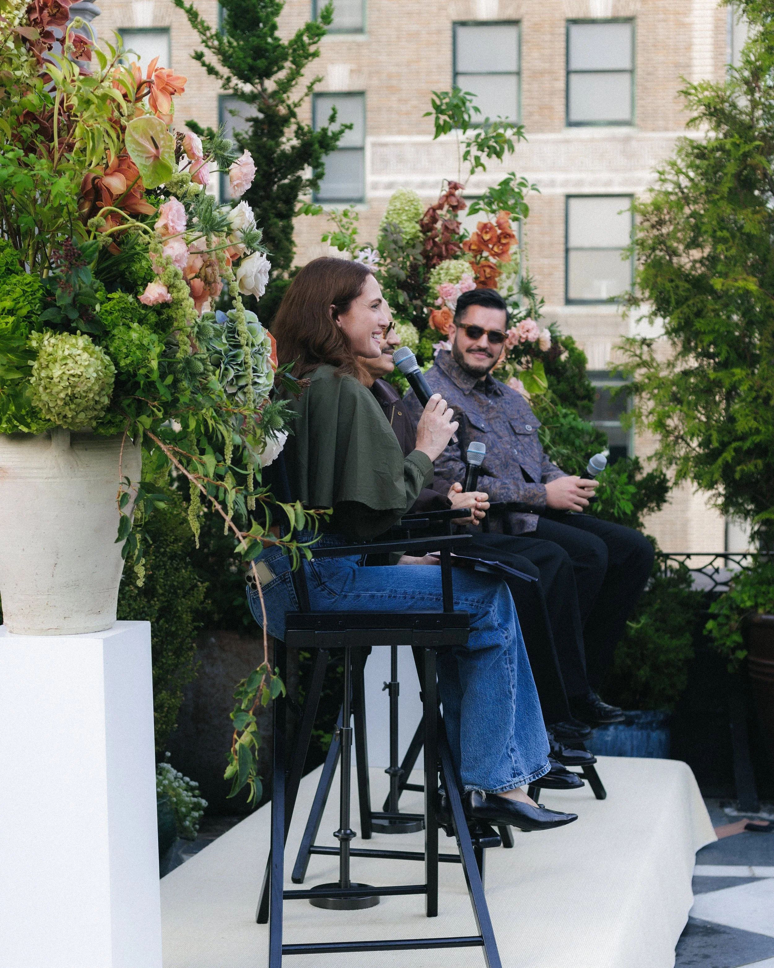 A woman speaking into a microphone outdoors at a panel discussion, with a man sitting beside her holding a microphone. There are lush plants and flowers around them, and a building with windows in the background.