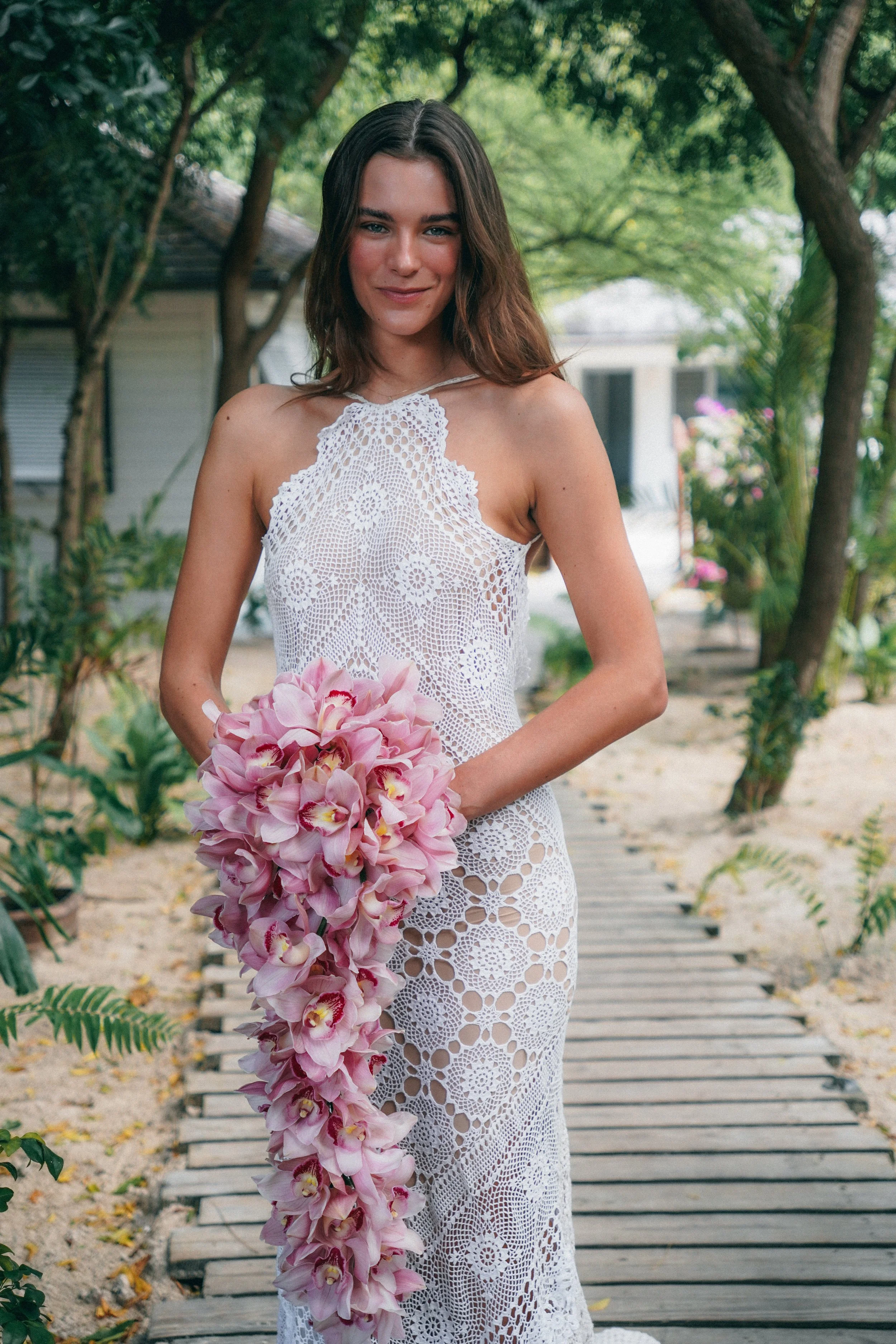 A woman in a white crochet dress holding a bouquet of pink orchids, standing on a wooden pathway surrounded by green trees.