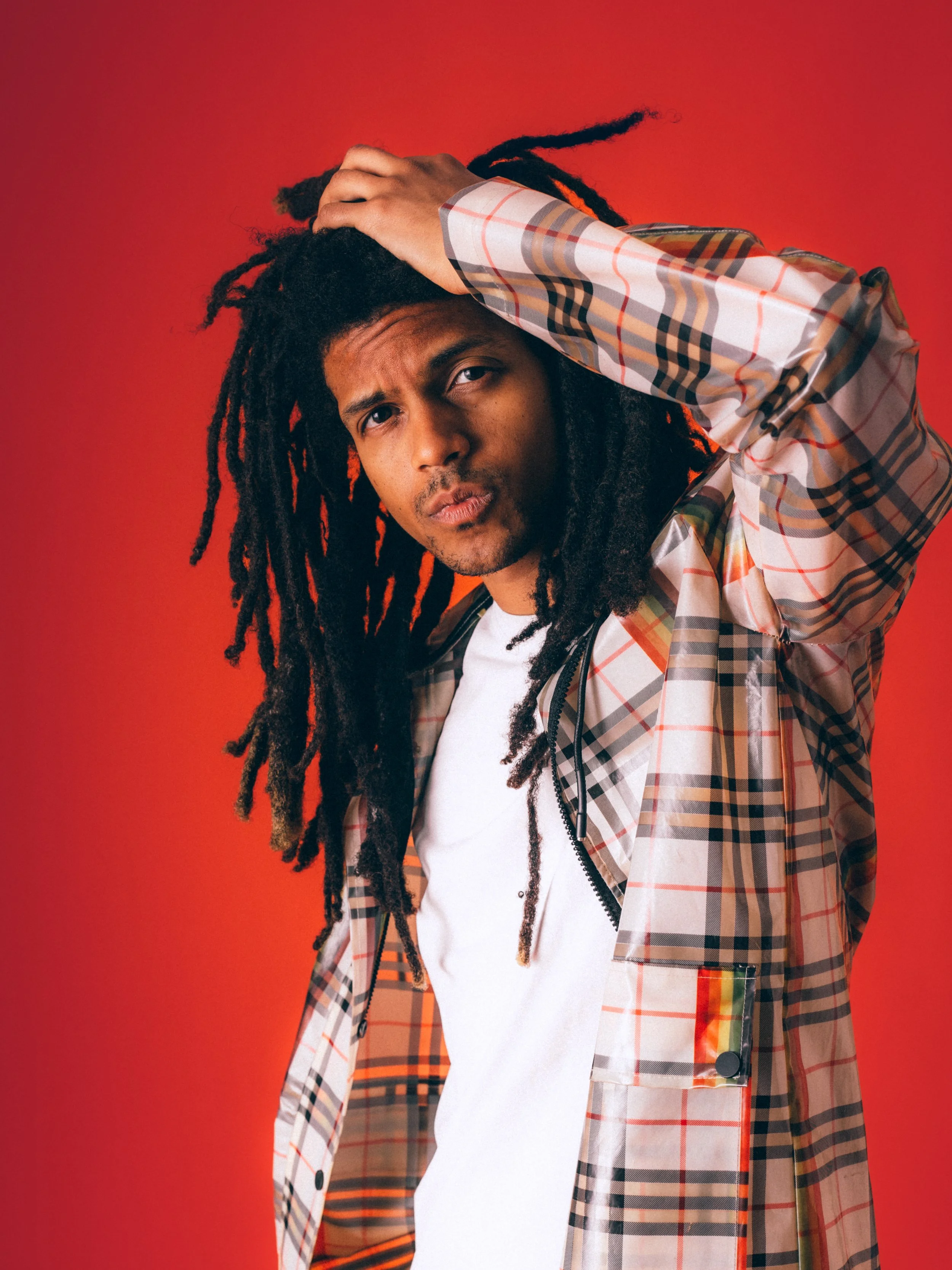A man with long dreadlocks and a serious expression, wearing a white t-shirt and a plaid jacket, holding his head with his hand against a red background.