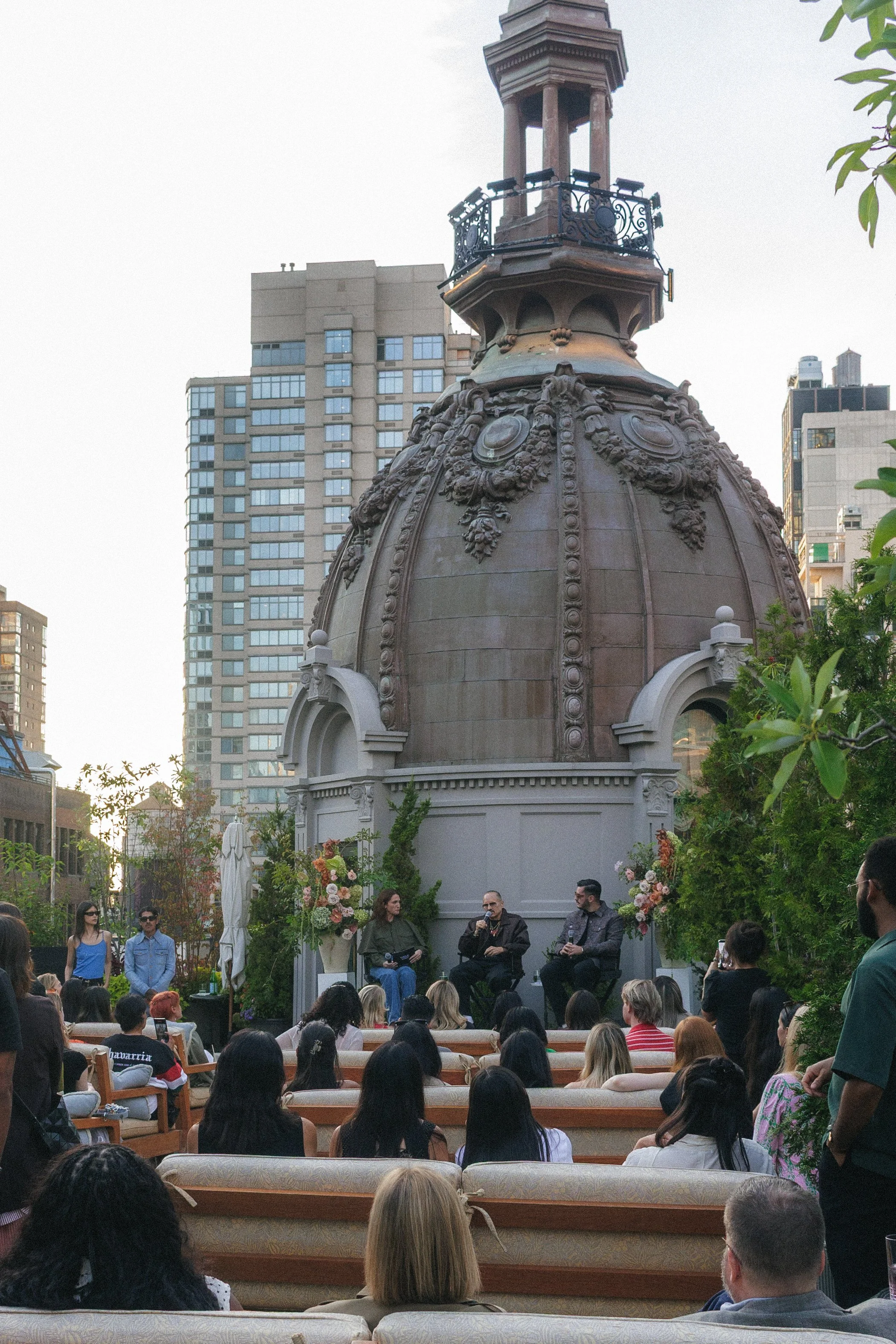 A panel discussion taking place outdoors on a small stage set against a historic building with a domed roof, surrounded by an audience seated on benches. The stage features three speakers and floral decorations.
