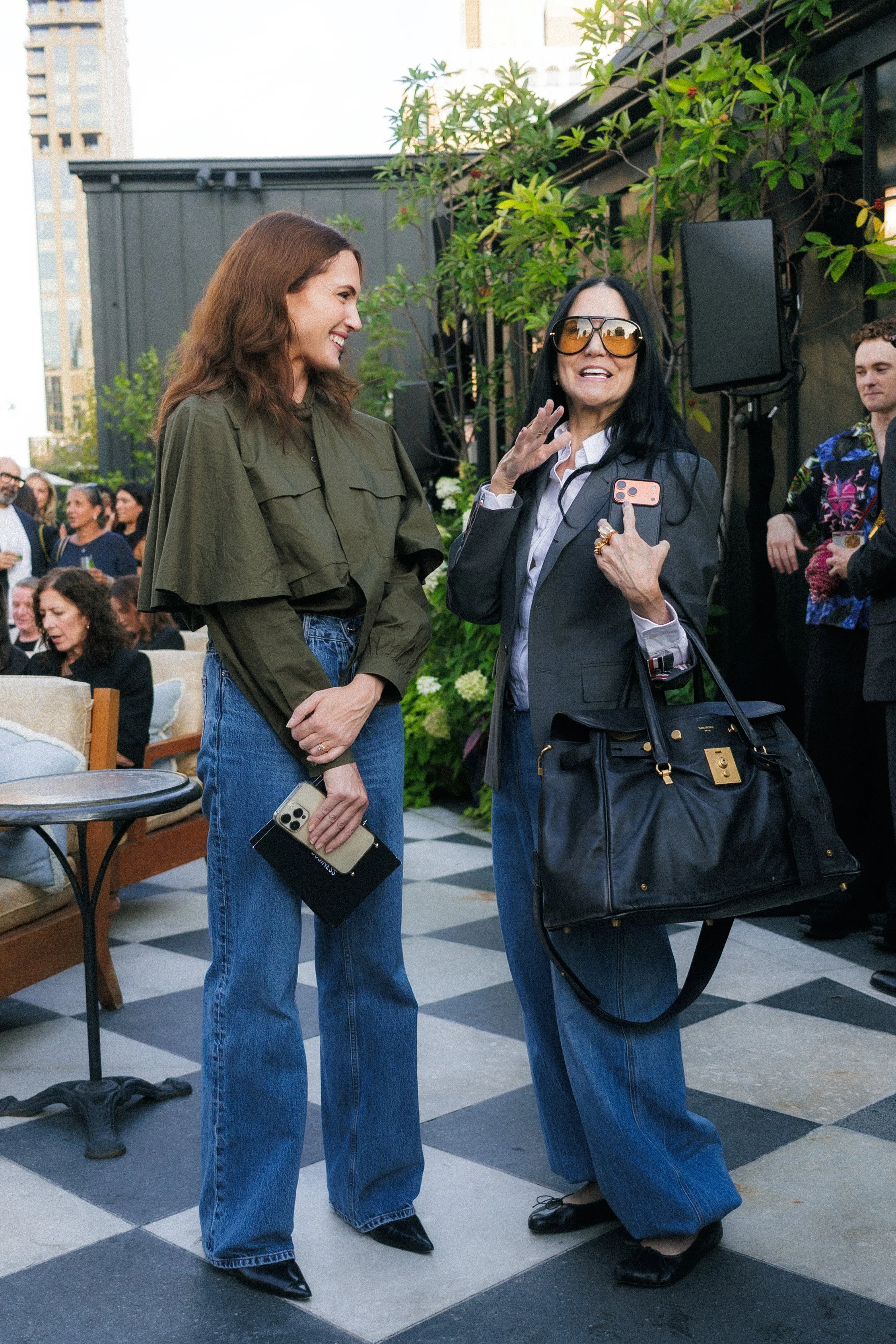 Two women are having a conversation at an outdoor event. One woman with long hair and glasses covered with tint is speaking and holding a smartphone, while the other woman with brown hair and a green jacket is smiling and listening. There are other p