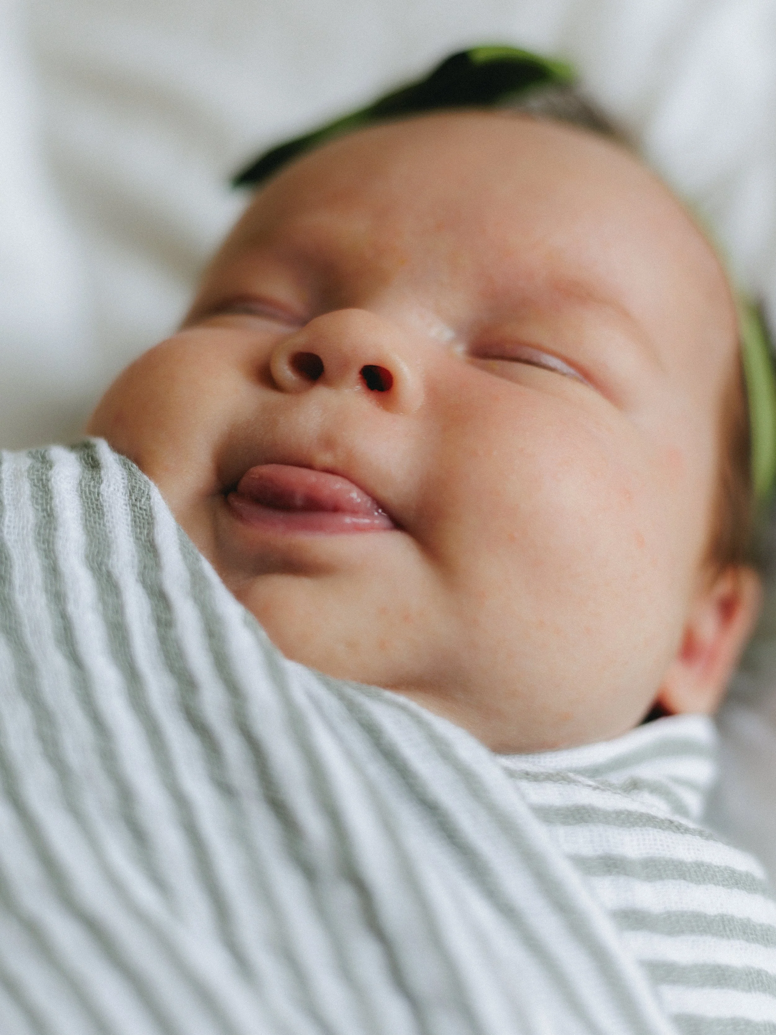 Close-up of a sleeping baby's face, showing the nose, closed eyes, and part of the mouth, with a hand in the foreground and a striped shirt.