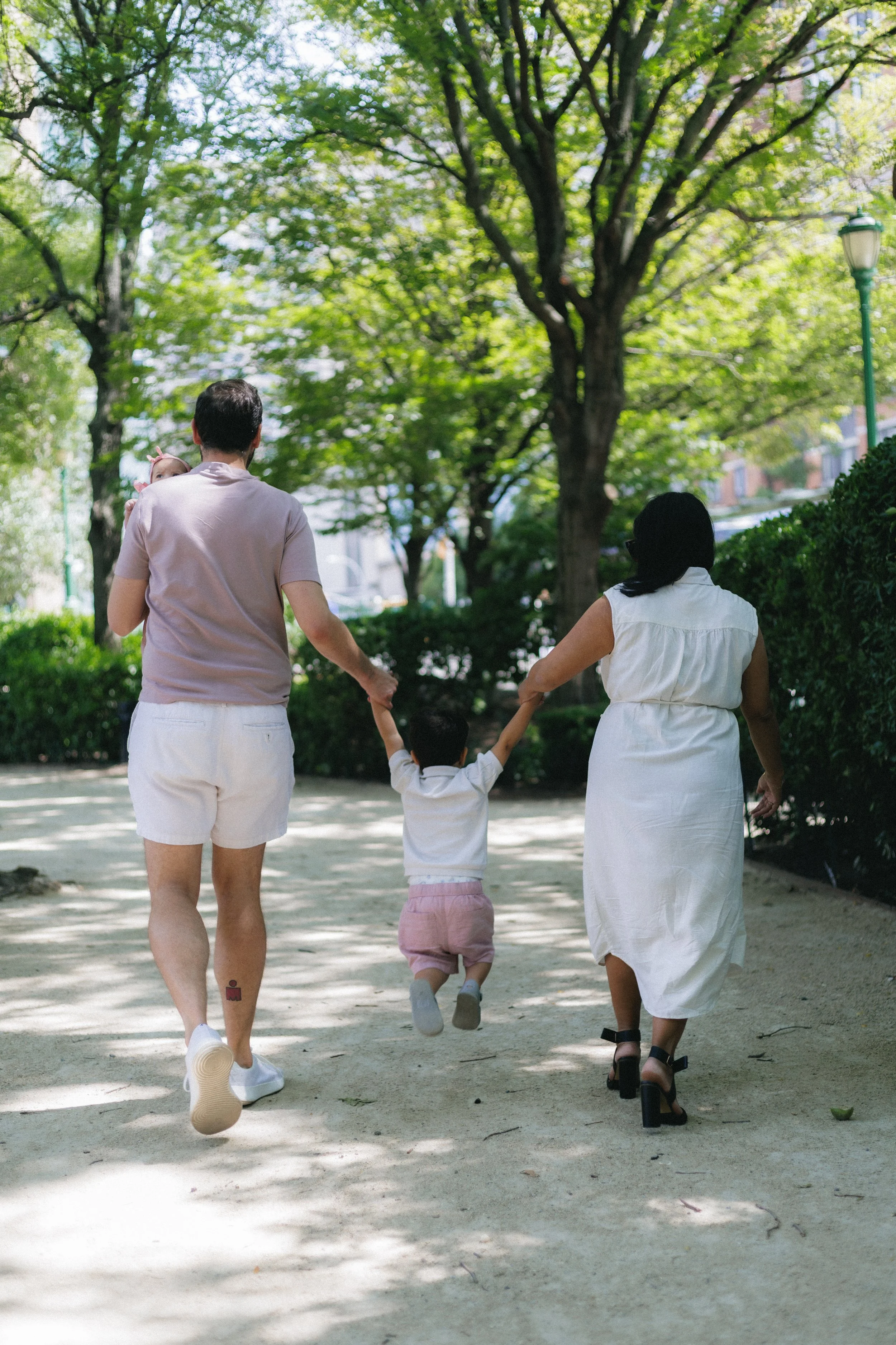 A family walking in a park on a sunny day, seen from behind. The family includes a man holding a child's hand, a woman, and a young child in the middle. The man is wearing a light-colored t-shirt and white shorts, the woman a white dress, and the chi