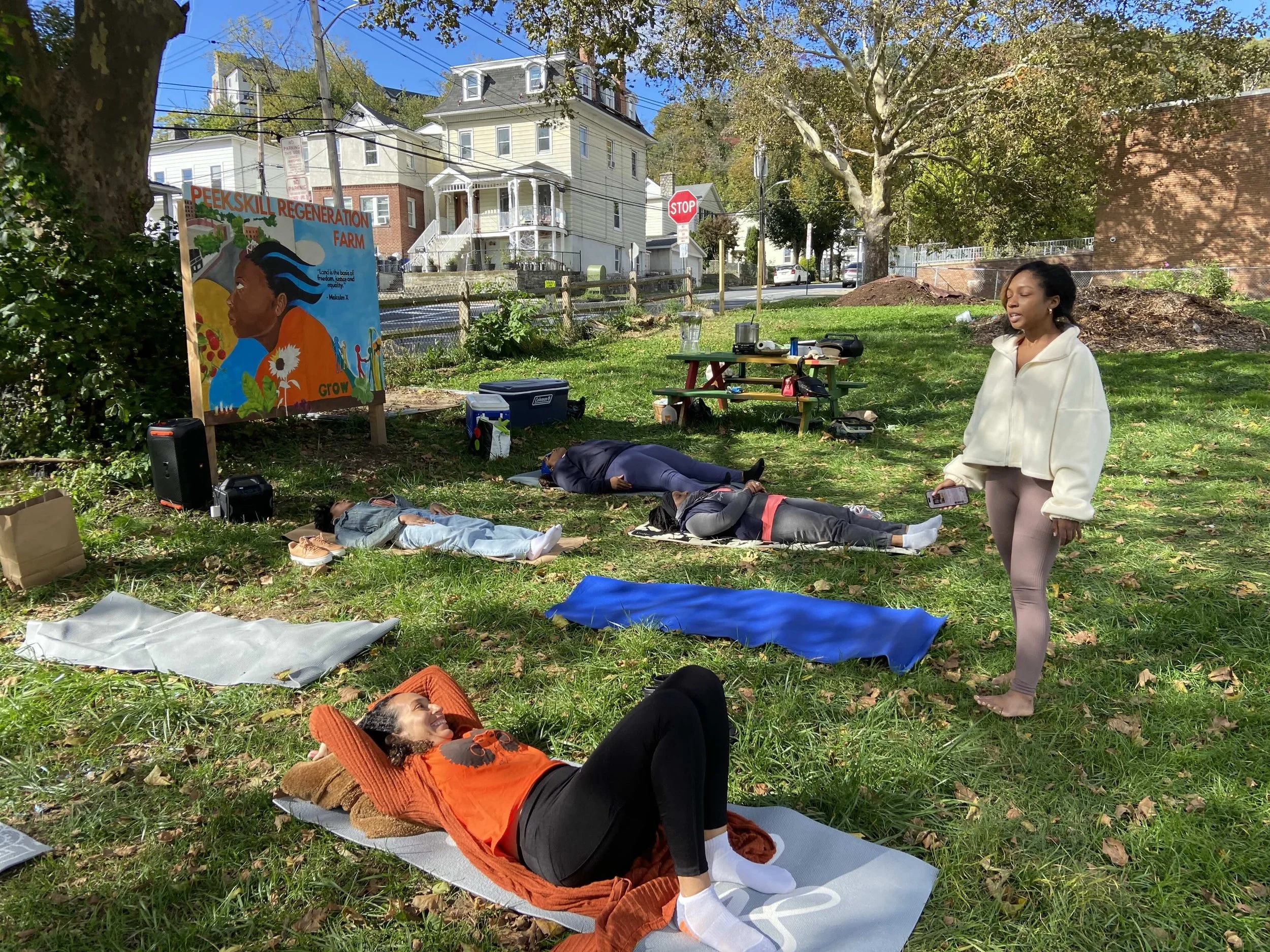 Group of people participating in an outdoor meditation session in a park, lying on yoga mats with their eyes closed, sun shining, and colorful artwork in the background.