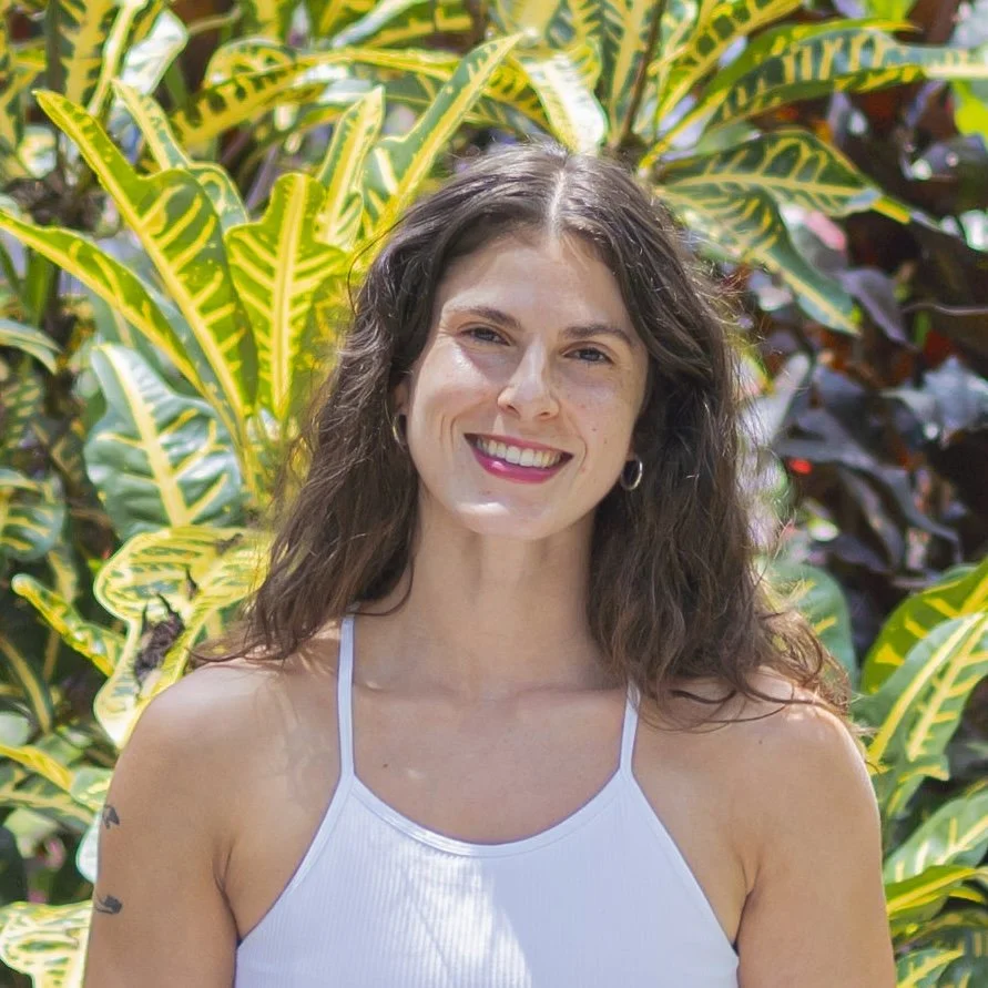 A woman smiling outdoors with green and yellow leafy plants behind her.