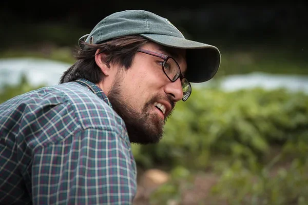A man with glasses and a beard wearing a green cap and plaid shirt outdoors with greenery in the background.