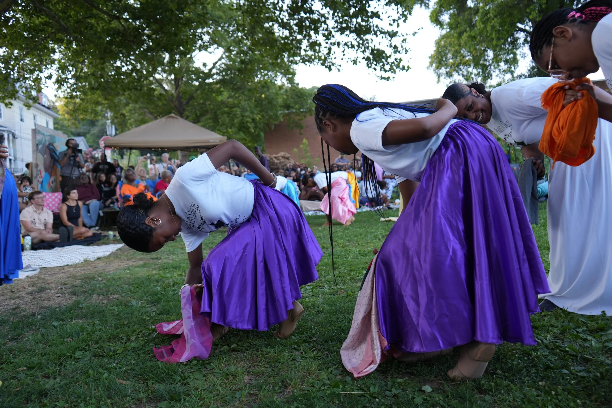Three women bowing during an outdoor performance, wearing purple skirts and white tops, with a crowd watching in the background under trees.