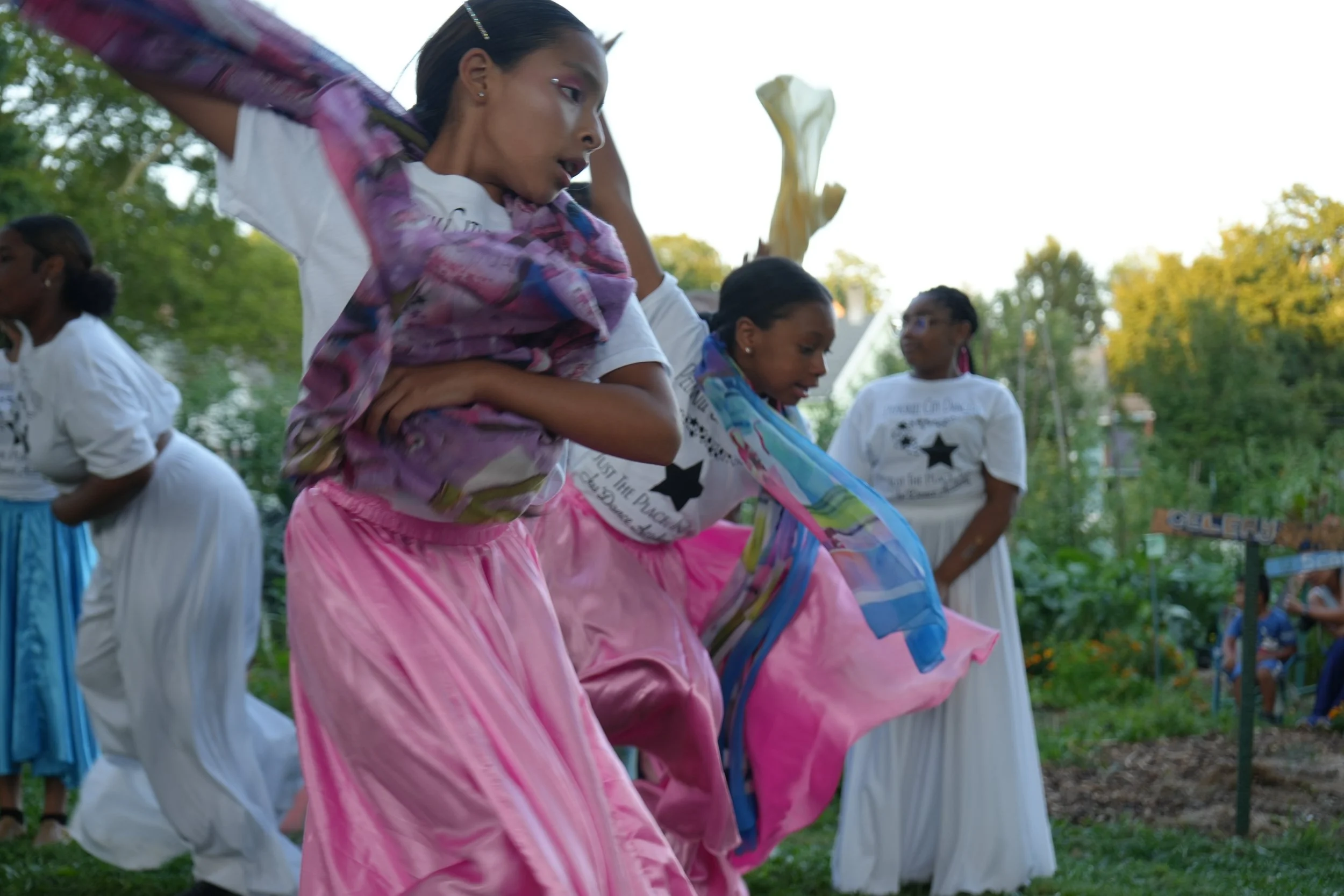 Young girls in colorful skirts dancing outdoors during a celebration or festival, with others watching in the background.