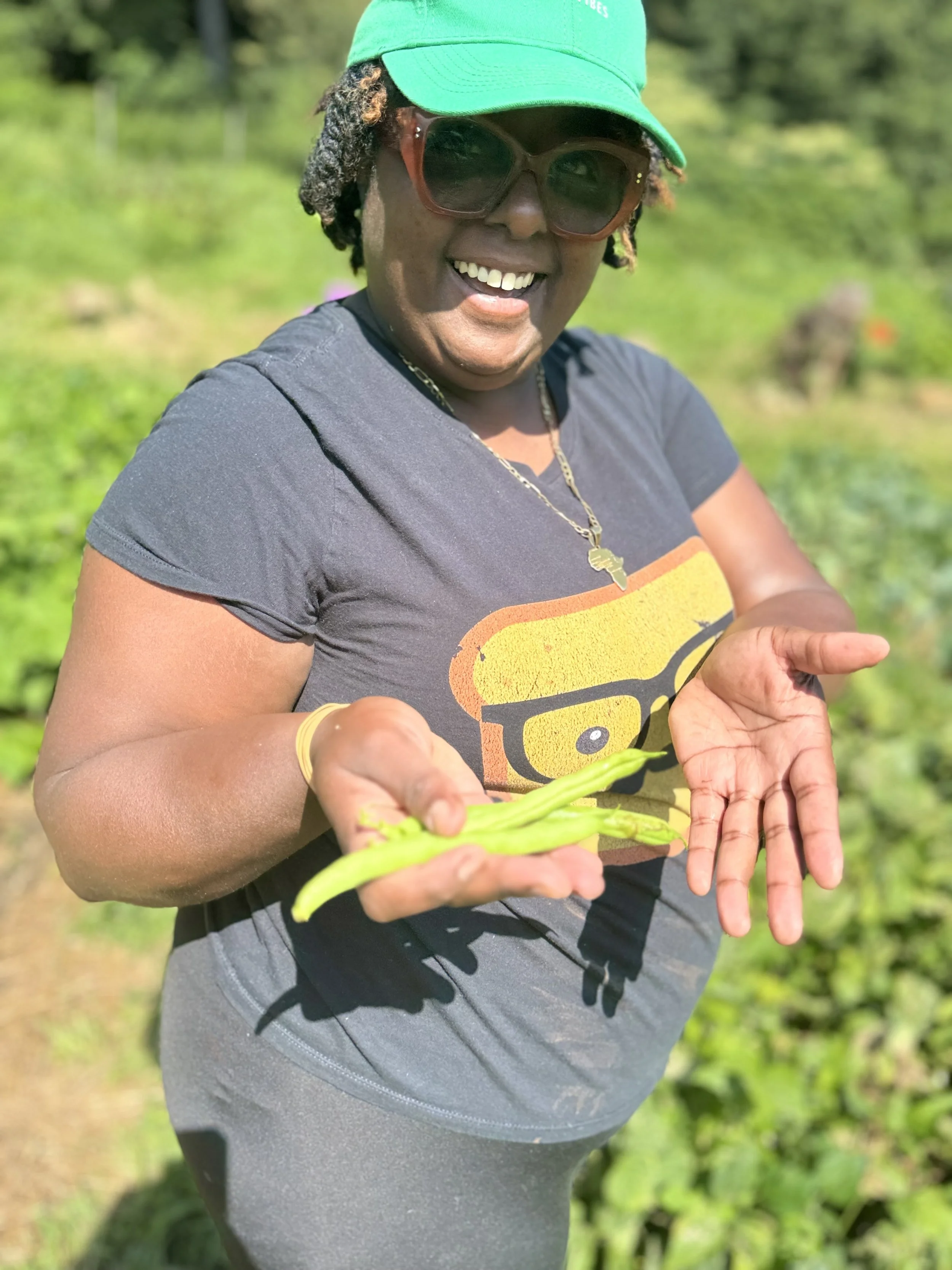 Smiling woman outdoors with sunglasses and green cap, holding a green chili pepper and showing her empty hands in a garden.
