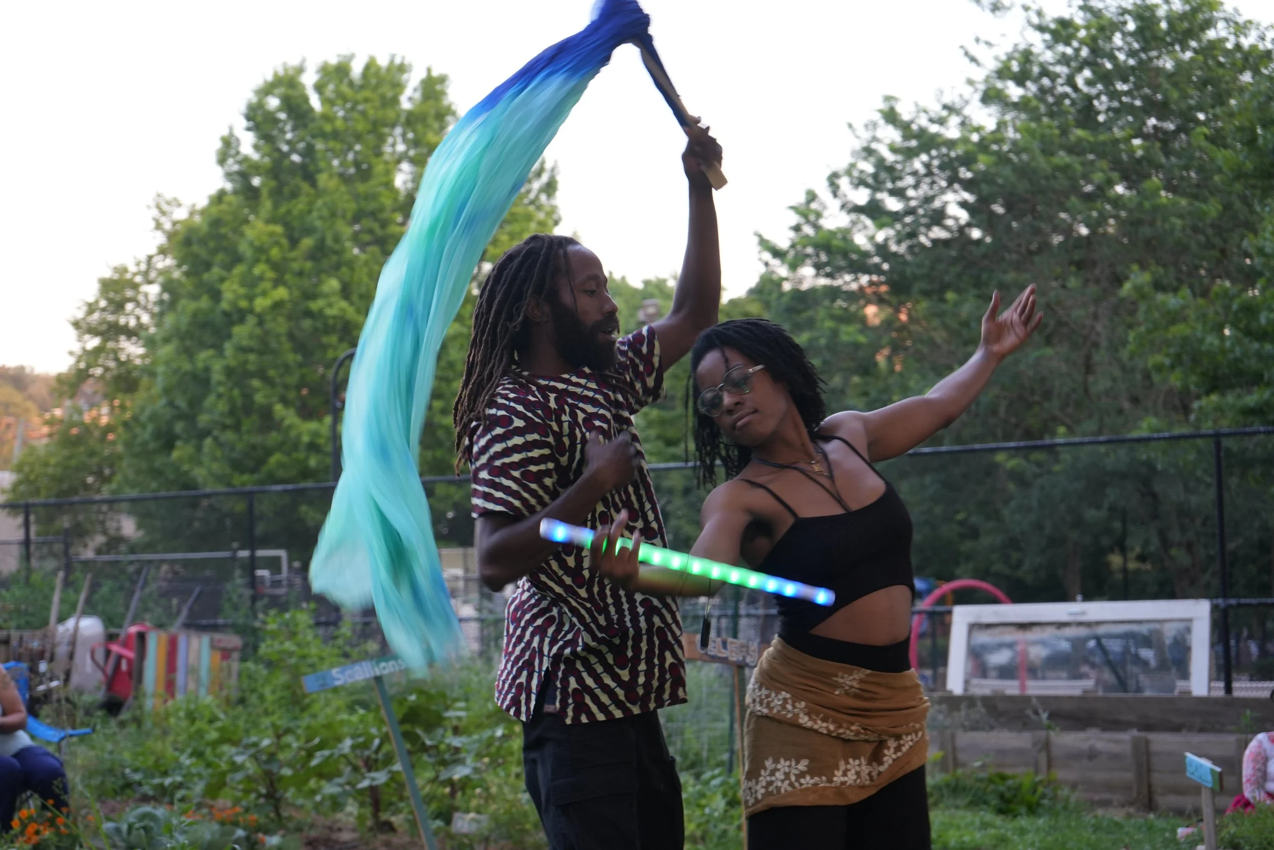 Two people dancing outdoors, one holding a lit LED poi with a blue and green fabric attached, surrounded by green trees and garden beds.