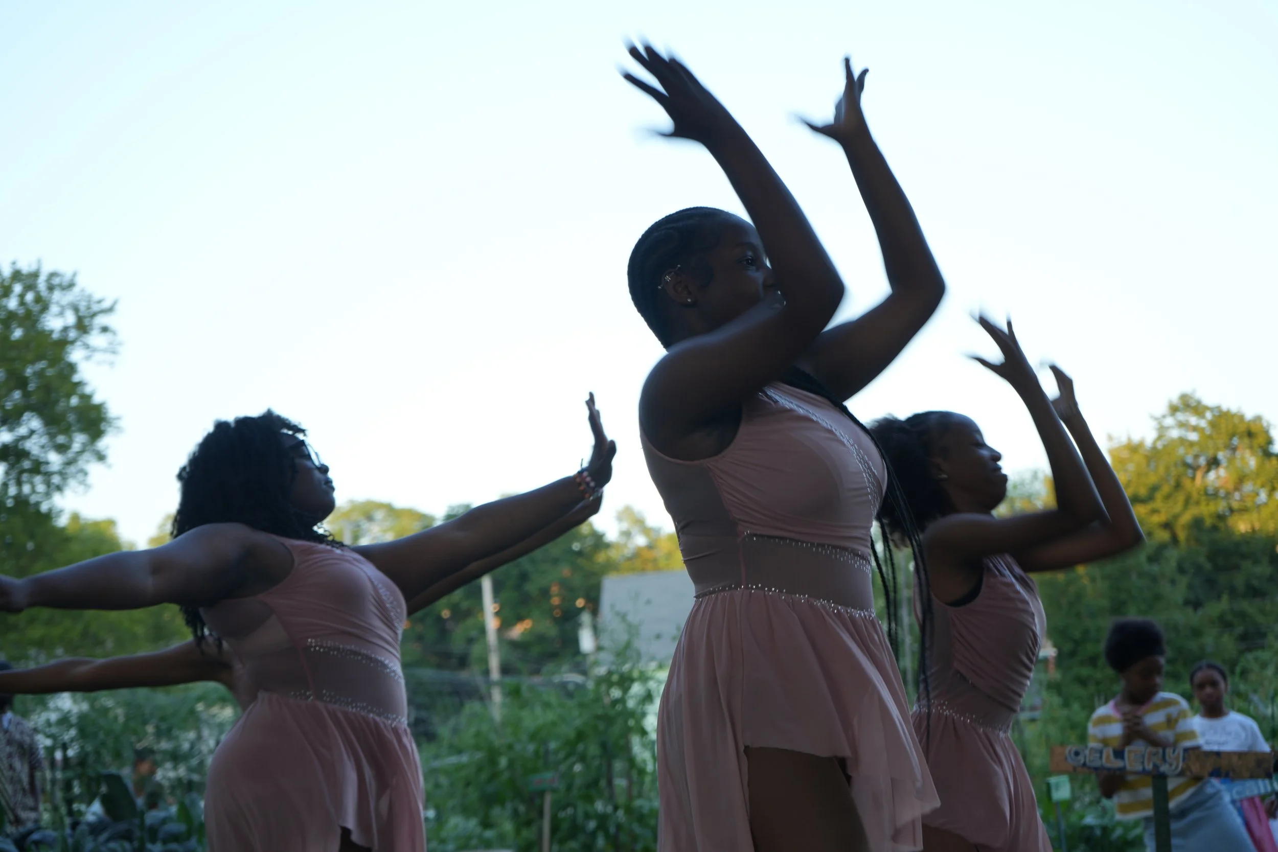 Three women in matching pink dresses dance outdoors with their arms raised during daytime.