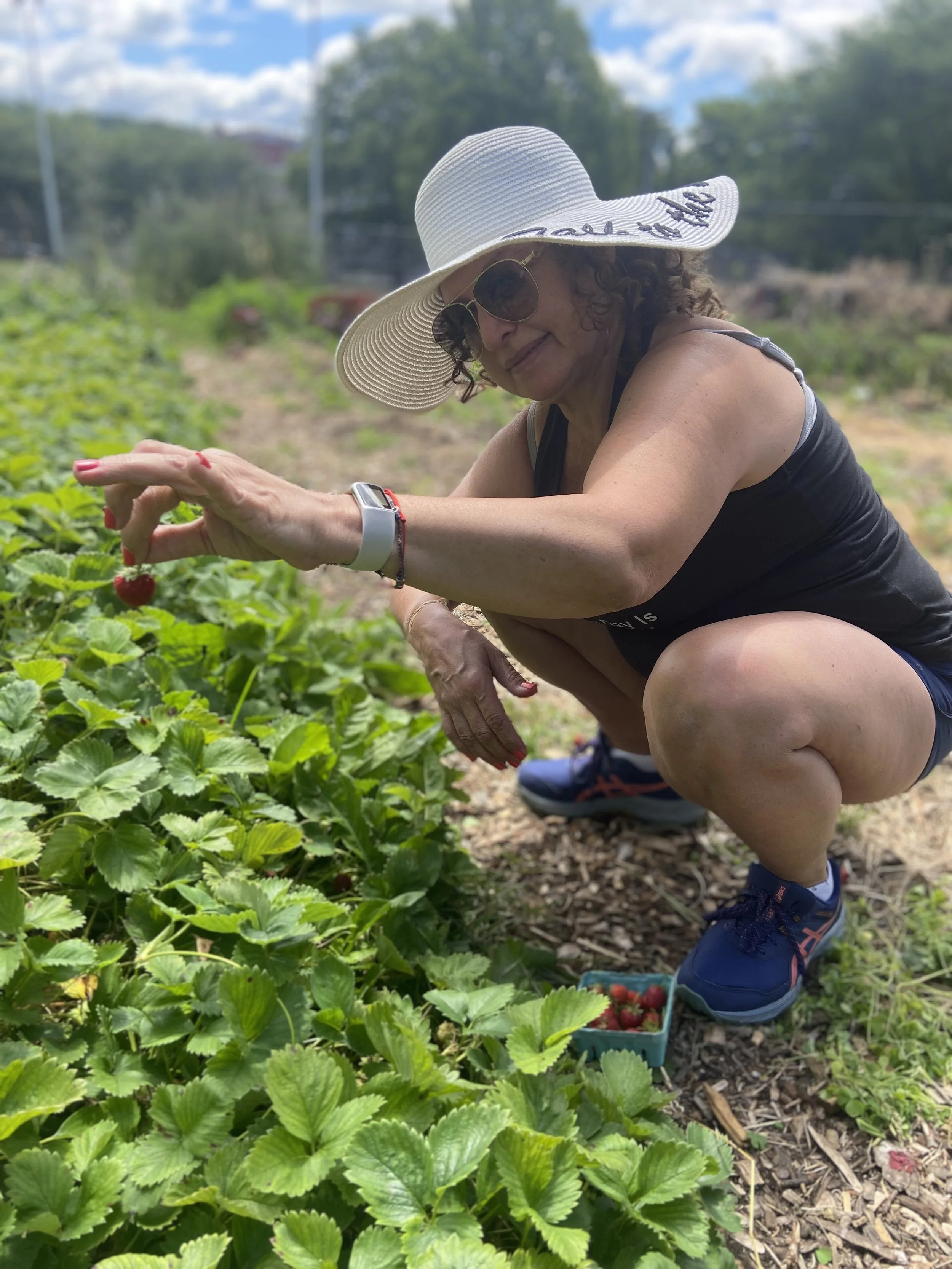A woman wearing a wide-brimmed hat, sunglasses, and athletic clothing kneels in a field to pick ripe strawberries from green plants. There is a box of strawberries on the ground beside her.