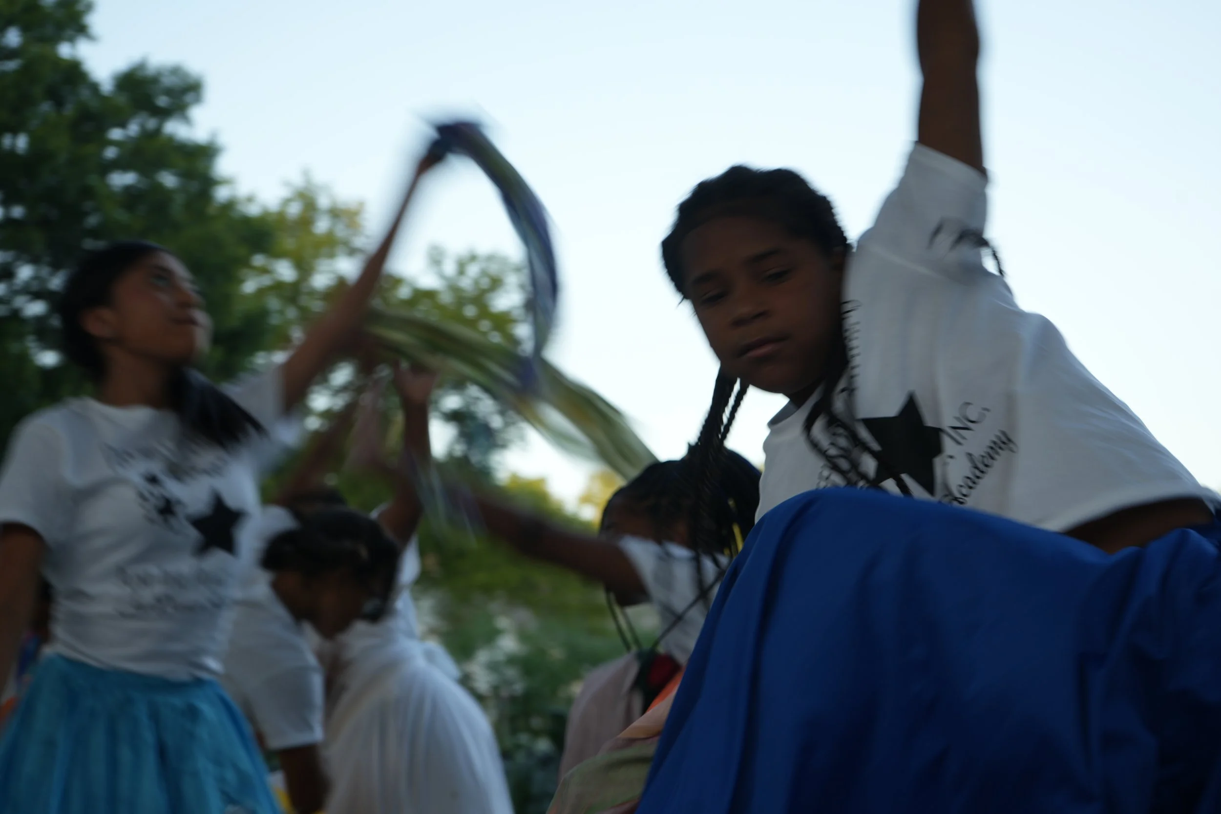 Group of children dancing outdoors during daytime, some with braided hair, wearing white t-shirts with black star logos and blue skirts or pants, with trees in the background.